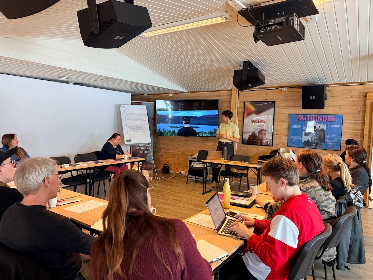 A presenter stands beside a screen showing a landscape film still while participants watch from tables arranged around the room