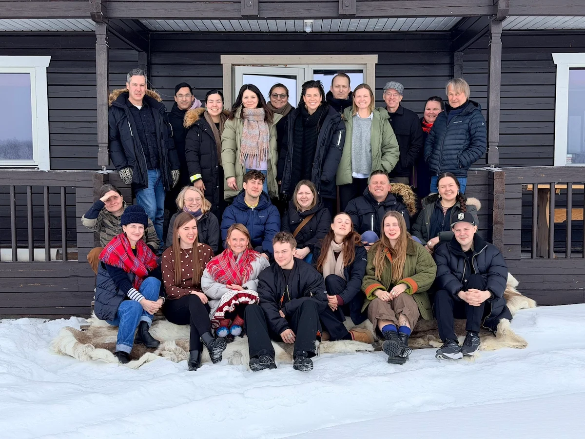 Group portrait of about twenty workshop participants on the snowy steps of a wooden lodge, some seated on reindeer hides