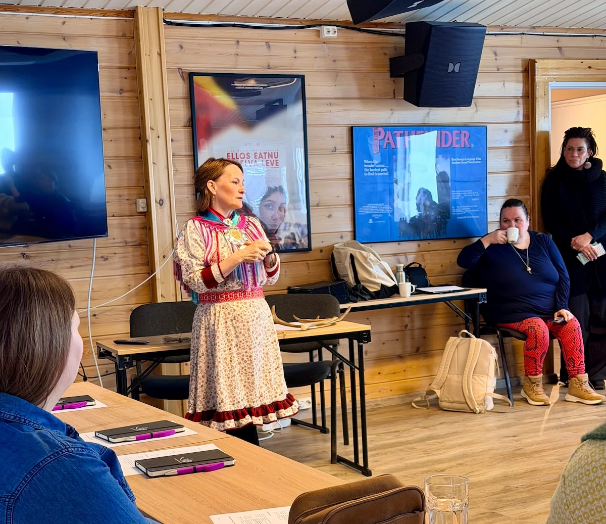 A woman in traditional Sámi dress speaks to a seated group in a wood-paneled room, with Sámi film posters on the wall behind her