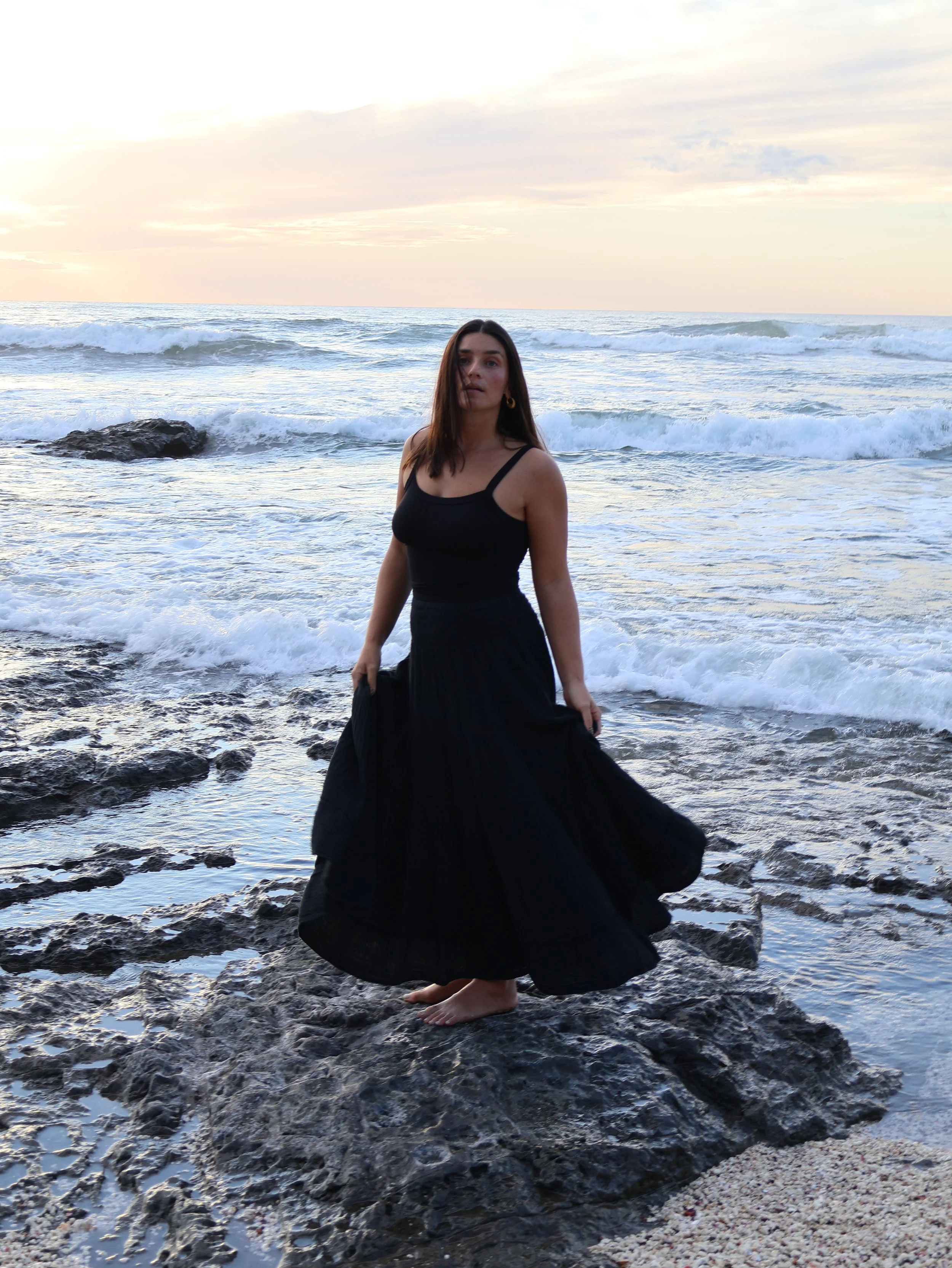 Young woman in black dress standing barefoot on rocks at the beach, ocean waves in background during sunset.