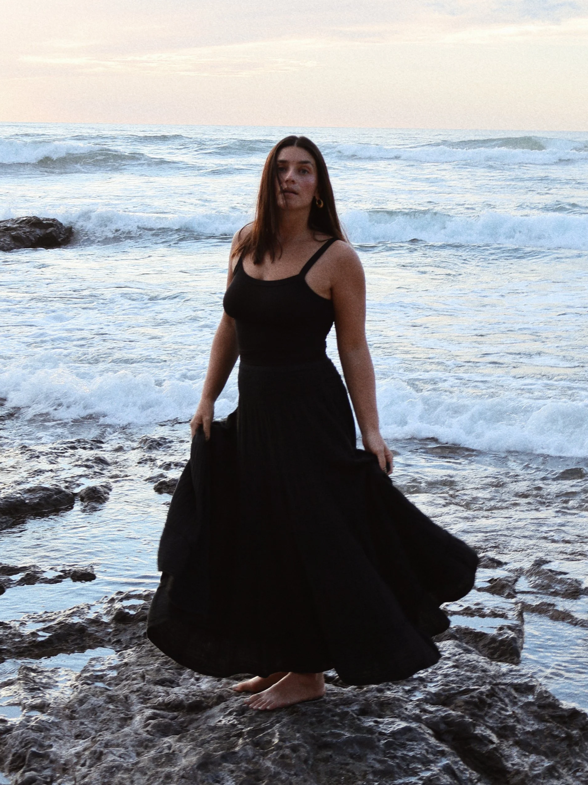 Young woman in a black dress standing barefoot on rocky shoreline with ocean waves and sky in the background.