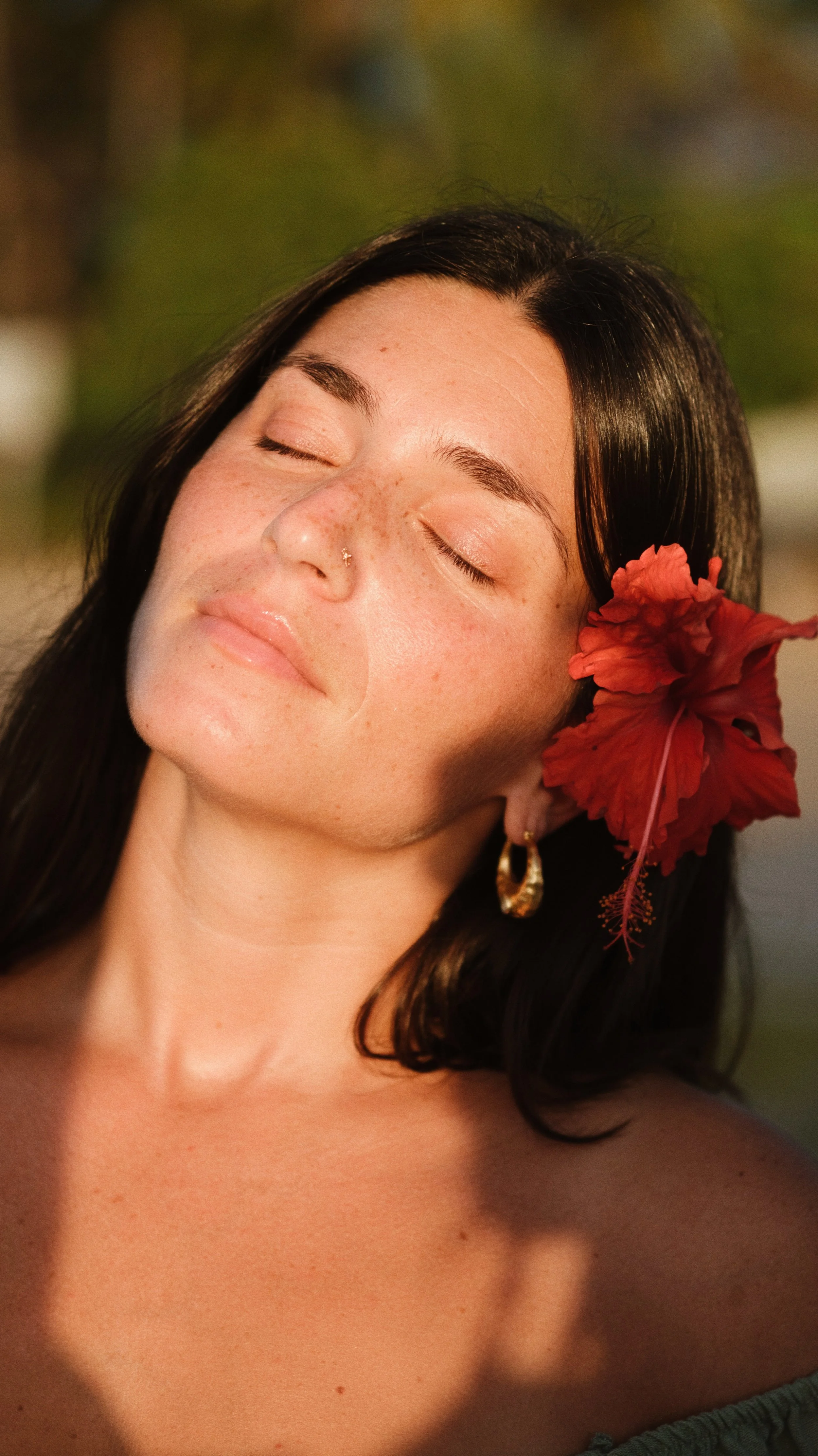 A woman with dark hair and freckles, with her eyes closed, wearing a gold hoop earring and a red flower in her hair.