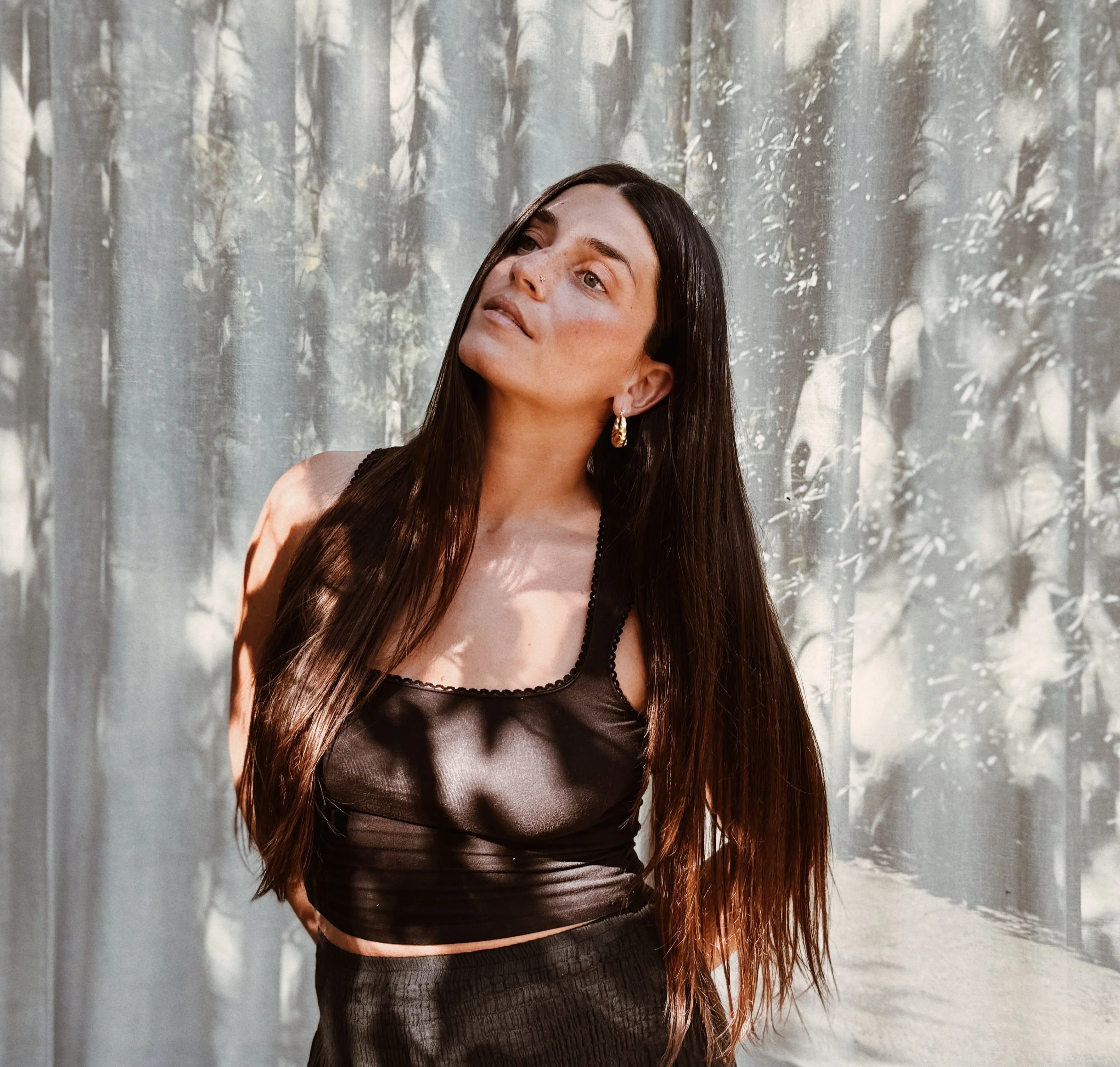 A woman with long, dark hair stands in front of a gray curtain with a forest pattern. She wears a black satin crop top and gold hoop earrings, looking slightly to the side with a thoughtful expression.