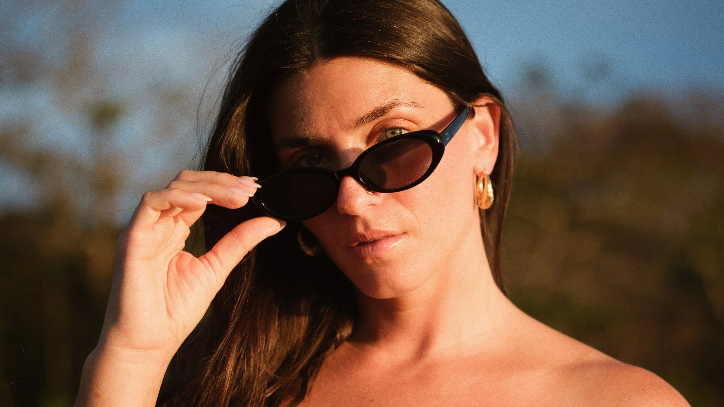 A woman with long brown hair, gold hoop earrings, and light makeup, looking at the camera while lowering sunglasses on her face outdoors during sunset.