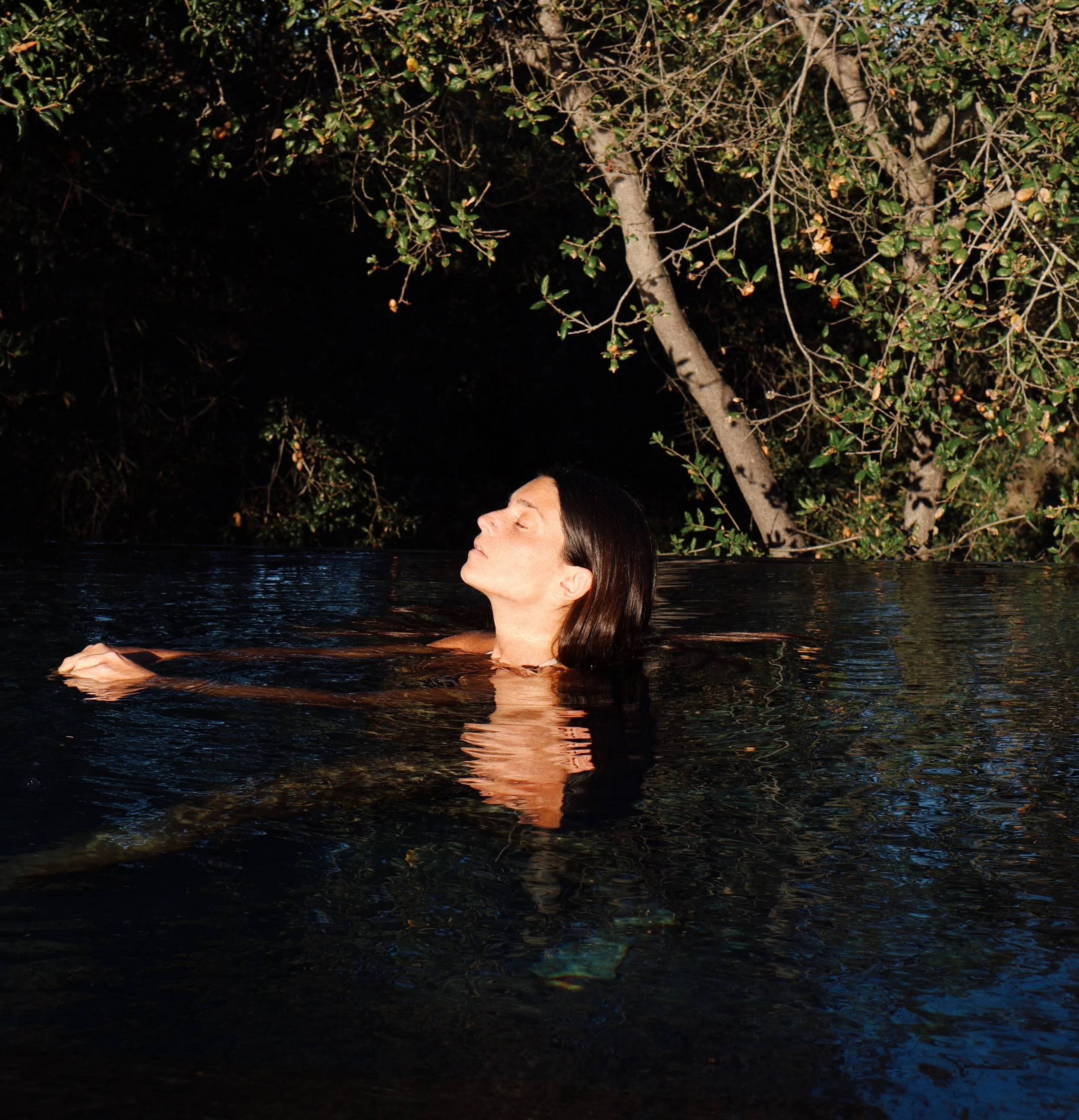 A woman is relaxing in a body of water with her eyes closed, surrounded by trees and foliage, at sunset or night.