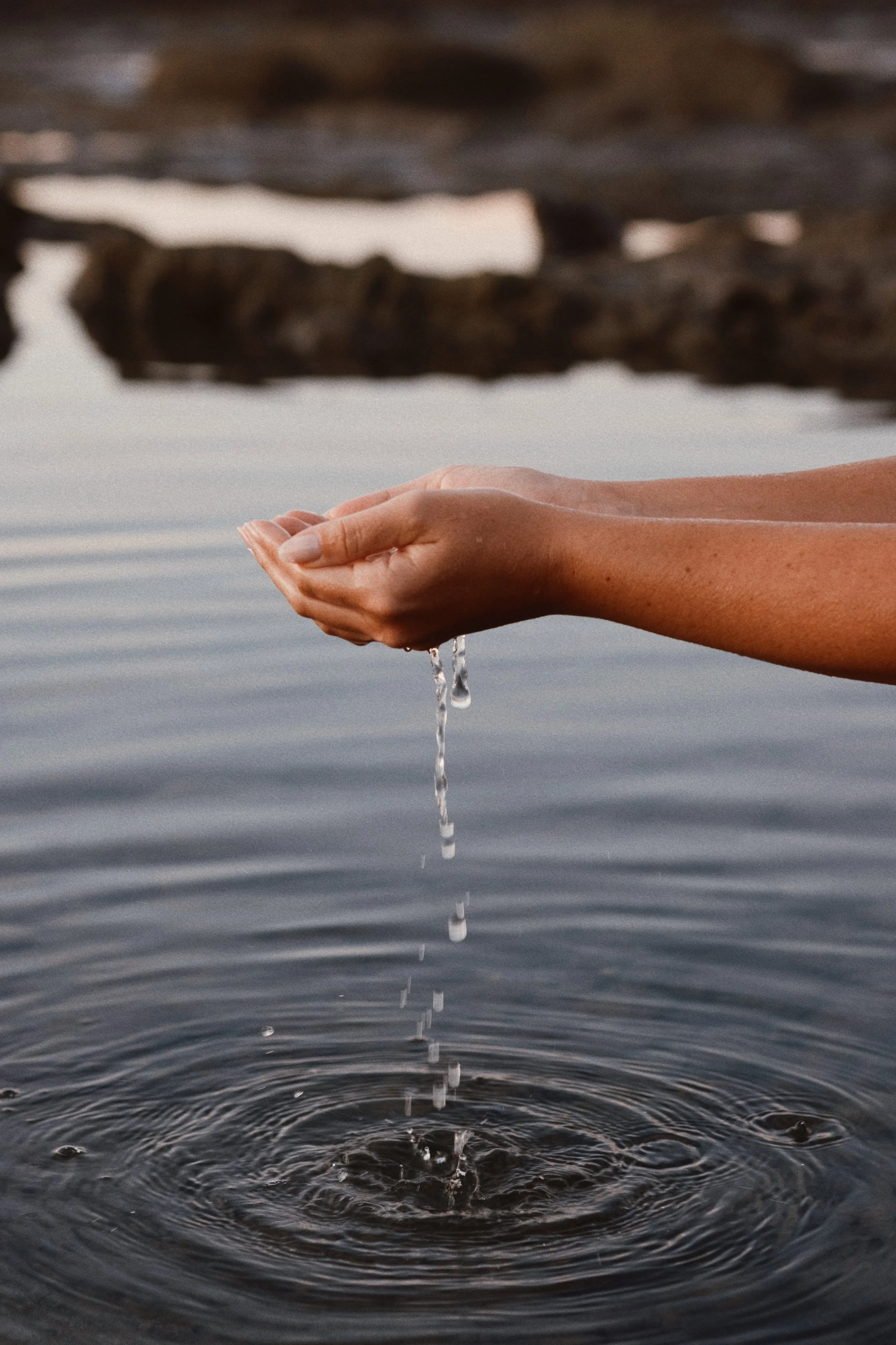 A close-up of hands cupping water above a body of water with small ripples, and rocks in the background.