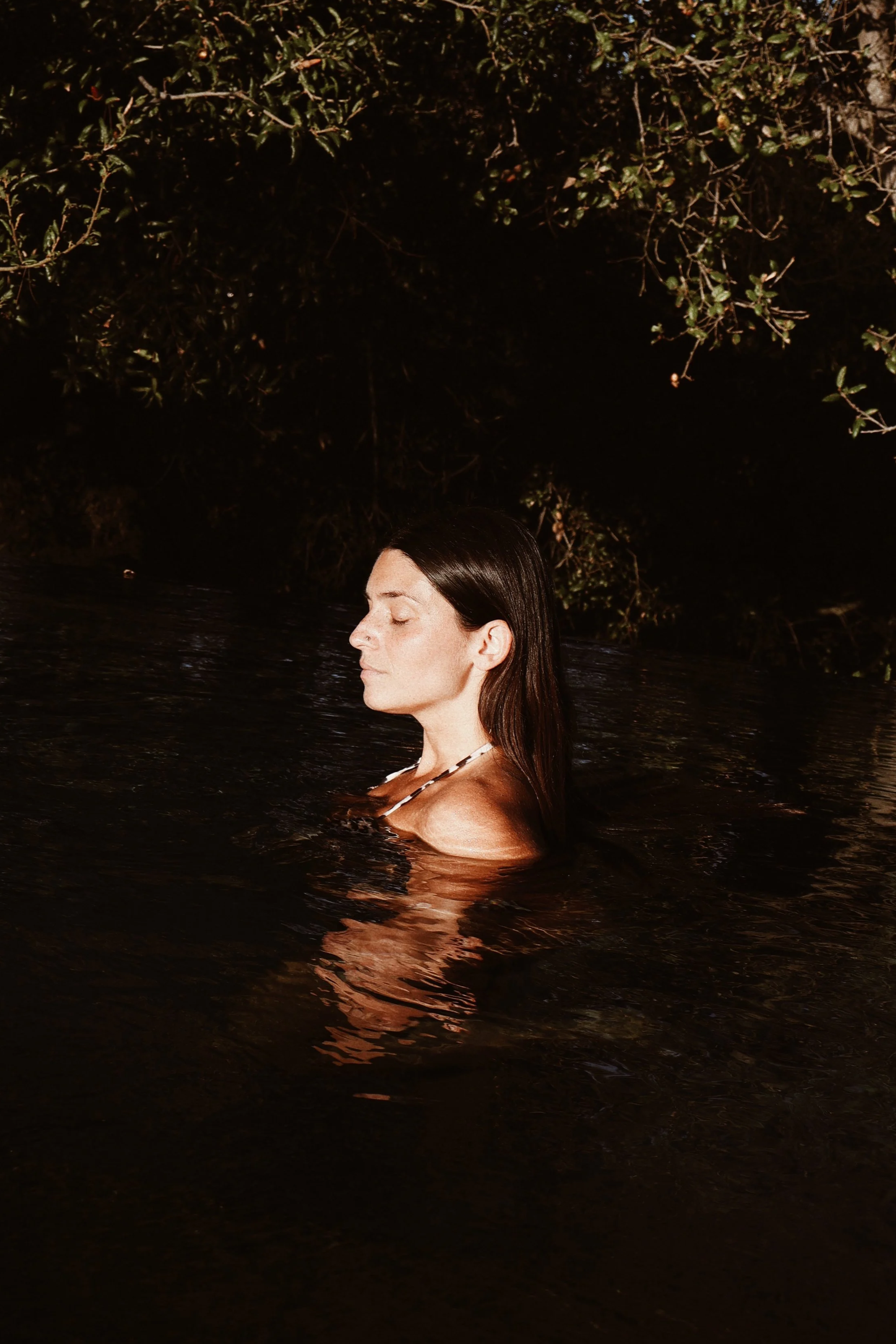 A woman with long dark hair submerged in water up to her shoulders at night, with her eyes closed and a peaceful expression, near bushes or trees.