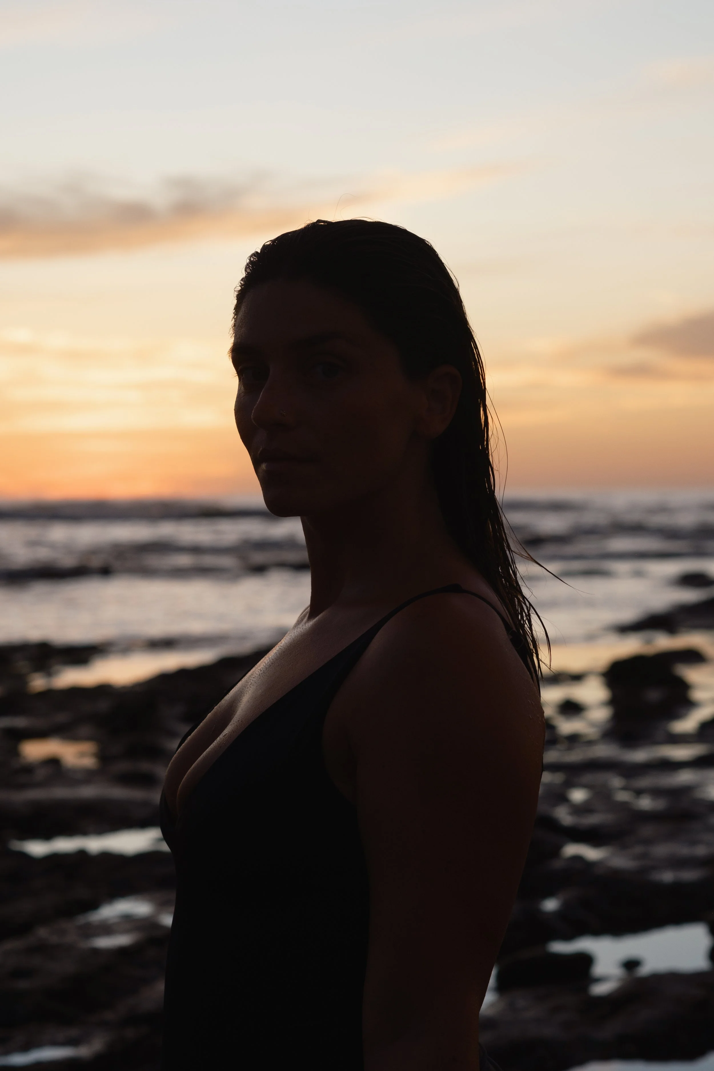 A person with wet hair standing at the beach during sunset, with the ocean and sky visible in the background.