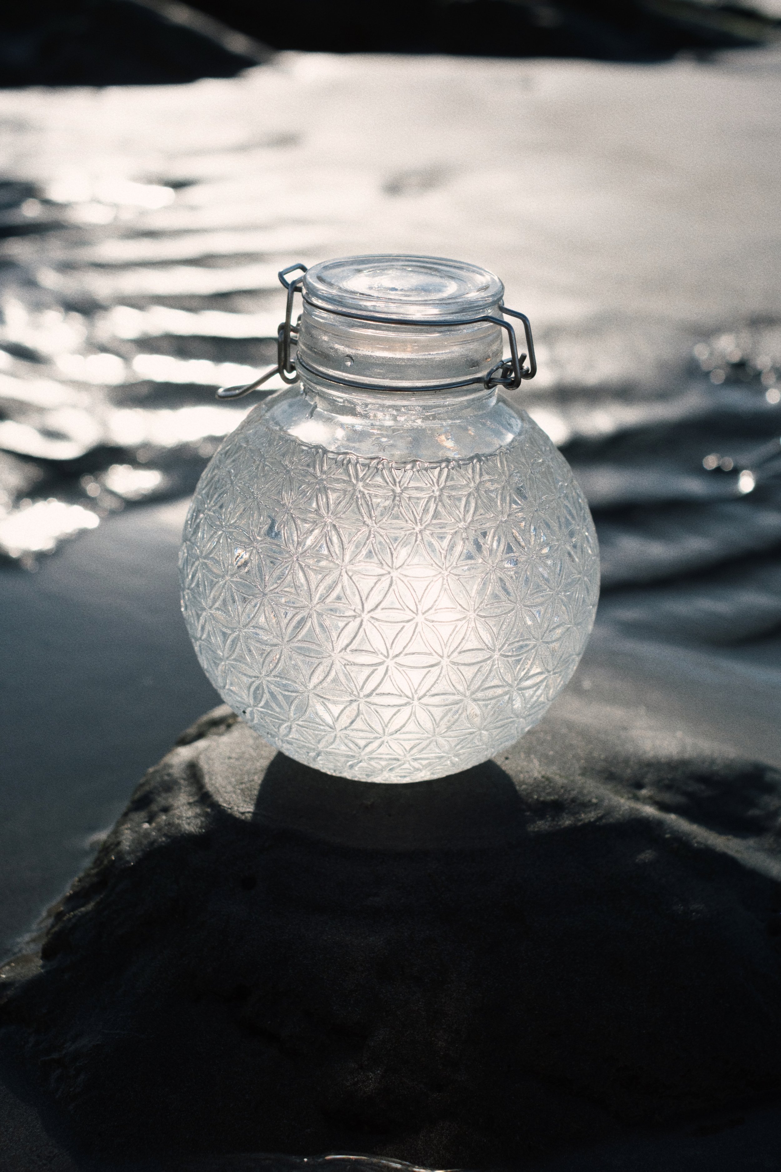 A clear glass jar with a textured geometric pattern, sitting on a rock near water, with some sunlight reflecting off the water's surface in the background.