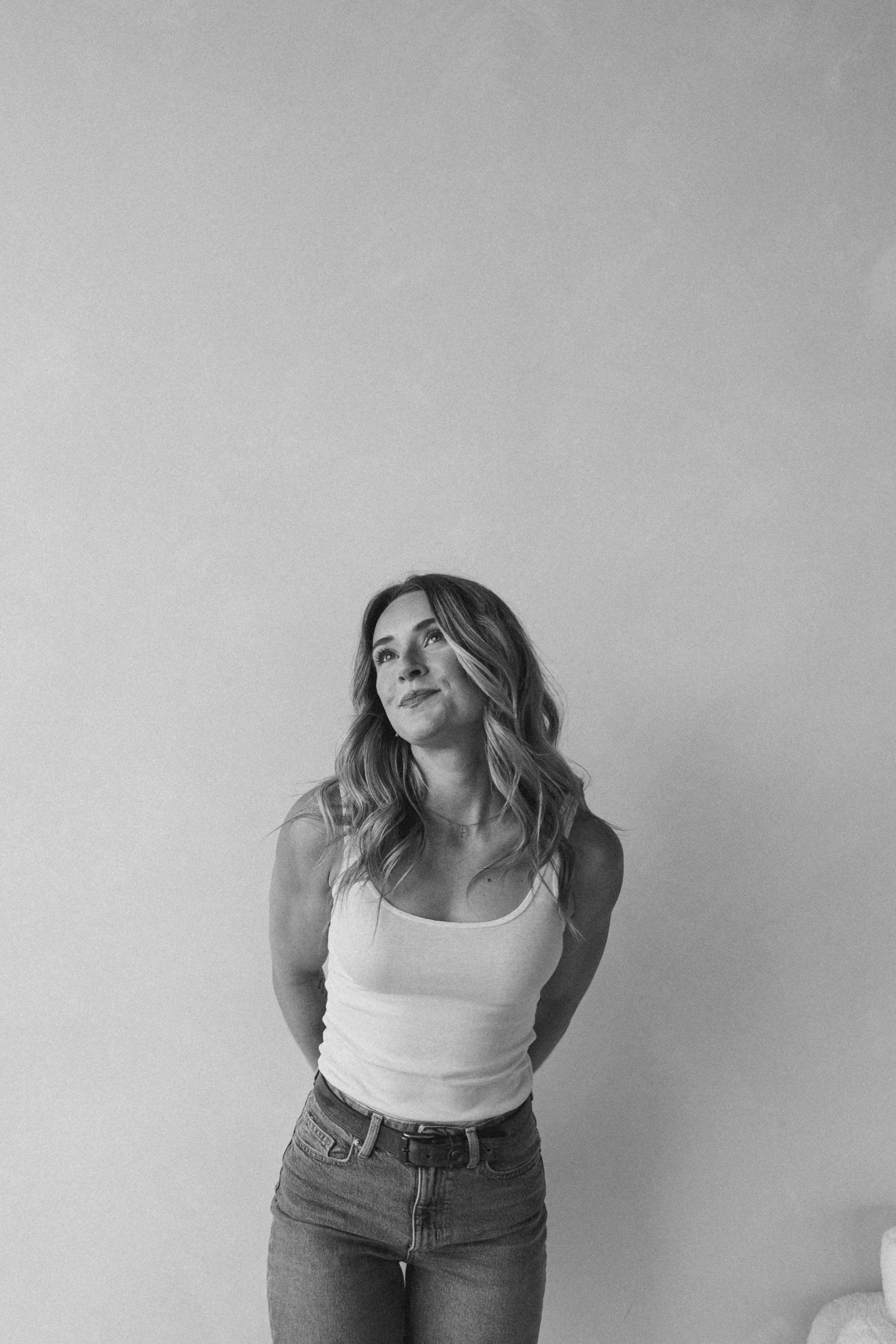 A woman with wavy hair wearing a tank top and jeans, standing against a plain wall, looking slightly upwards with a slight smile.