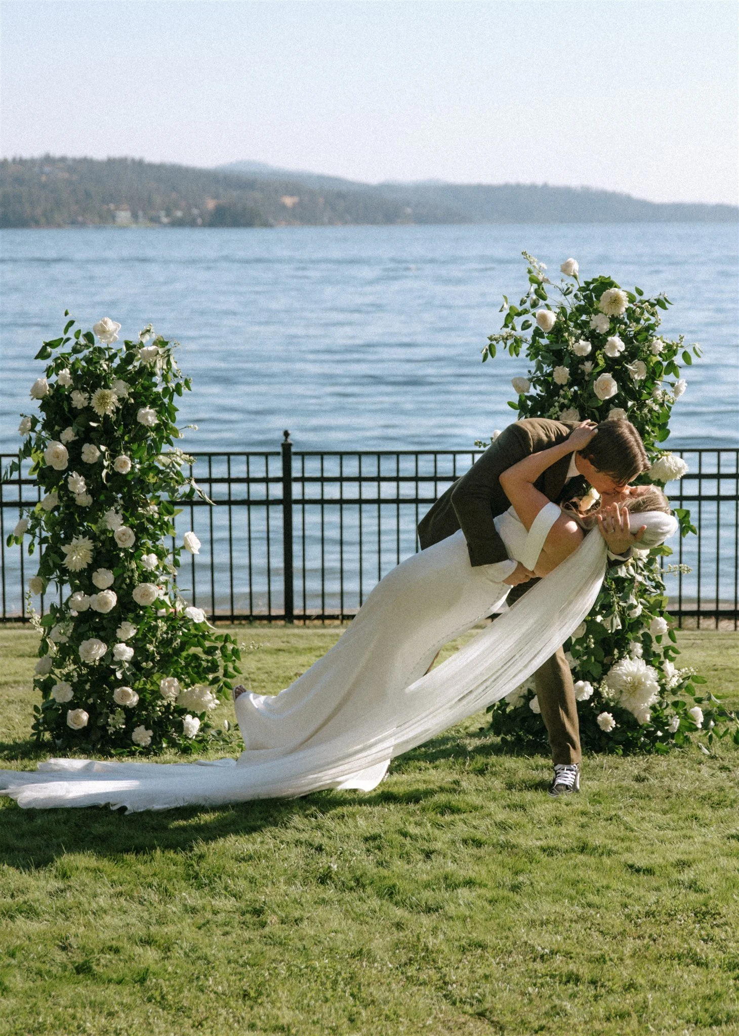 A newlywed couple sharing a romantic dance outdoors by a lake, surrounded by white floral arrangements and greenery.