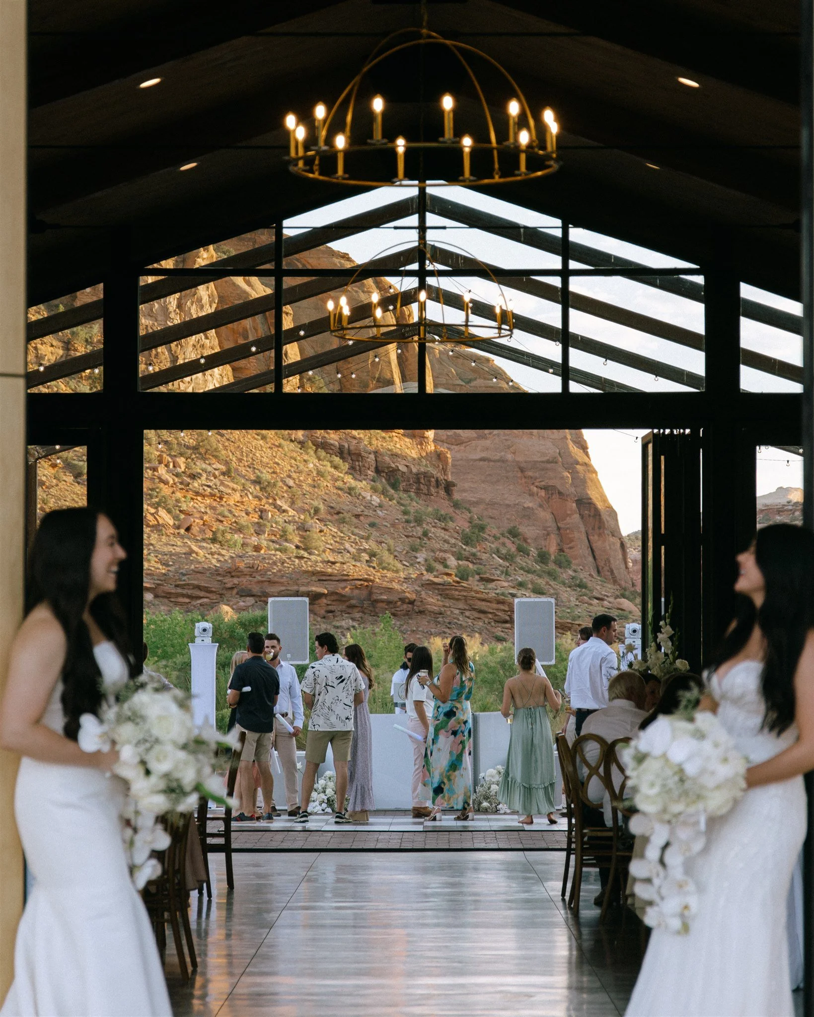 A wedding ceremony taking place inside a pavilion with open sides, with two brides in white dresses holding bouquets clustered together in the foreground, and guests and an officiant gathered near the outdoor altar with a scenic mountain backdrop.