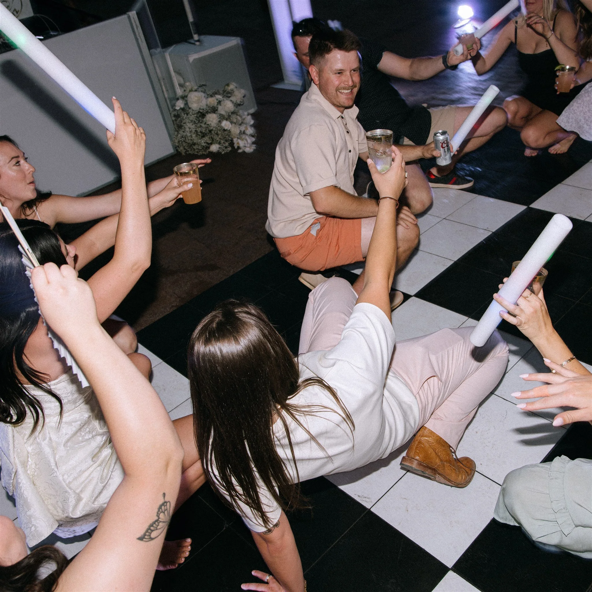 People gathered on the floor at a party, holding glow sticks and drinks, with some sitting and others lying down, in a lively and casual atmosphere.