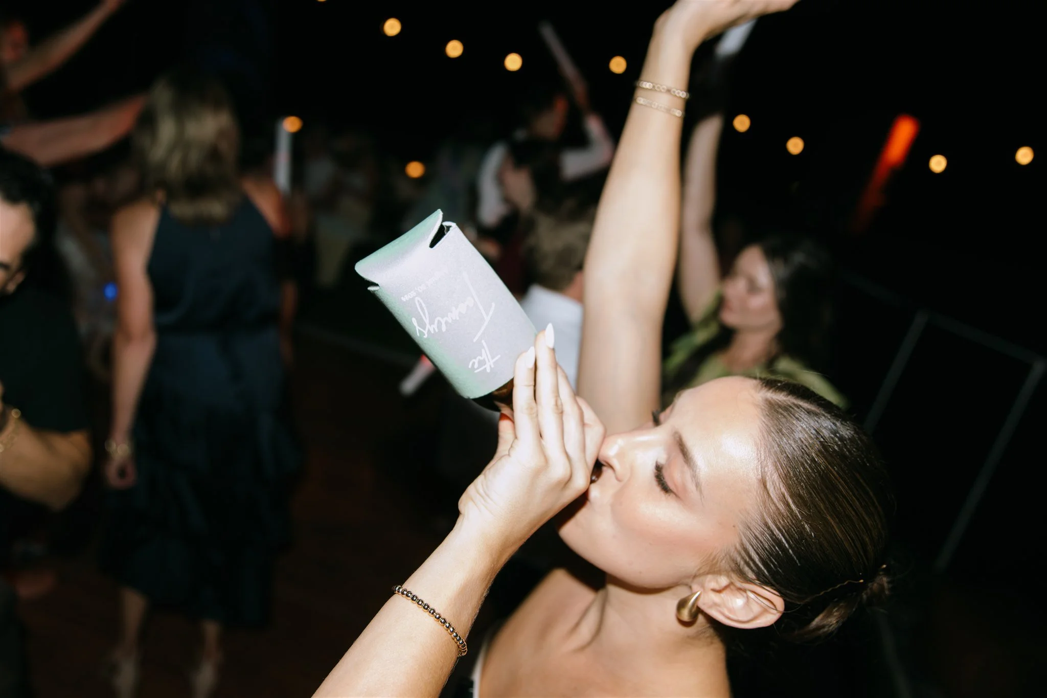 Woman kissing a champagne glass at a party with other people dancing in the background.