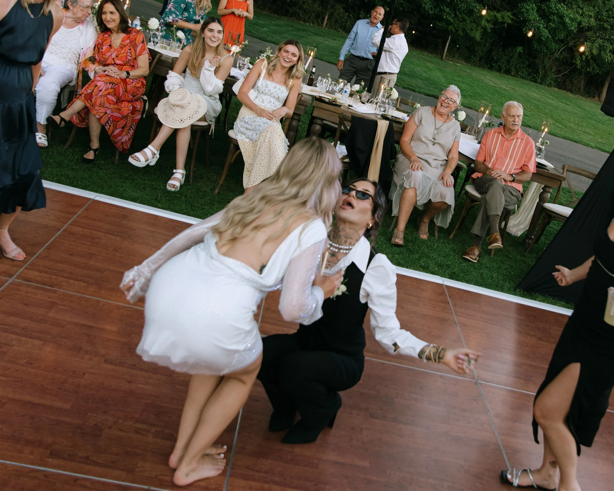 Two women are dancing closely on a wooden dance floor, surrounded by seated guests watching and smiling at an outdoor event.