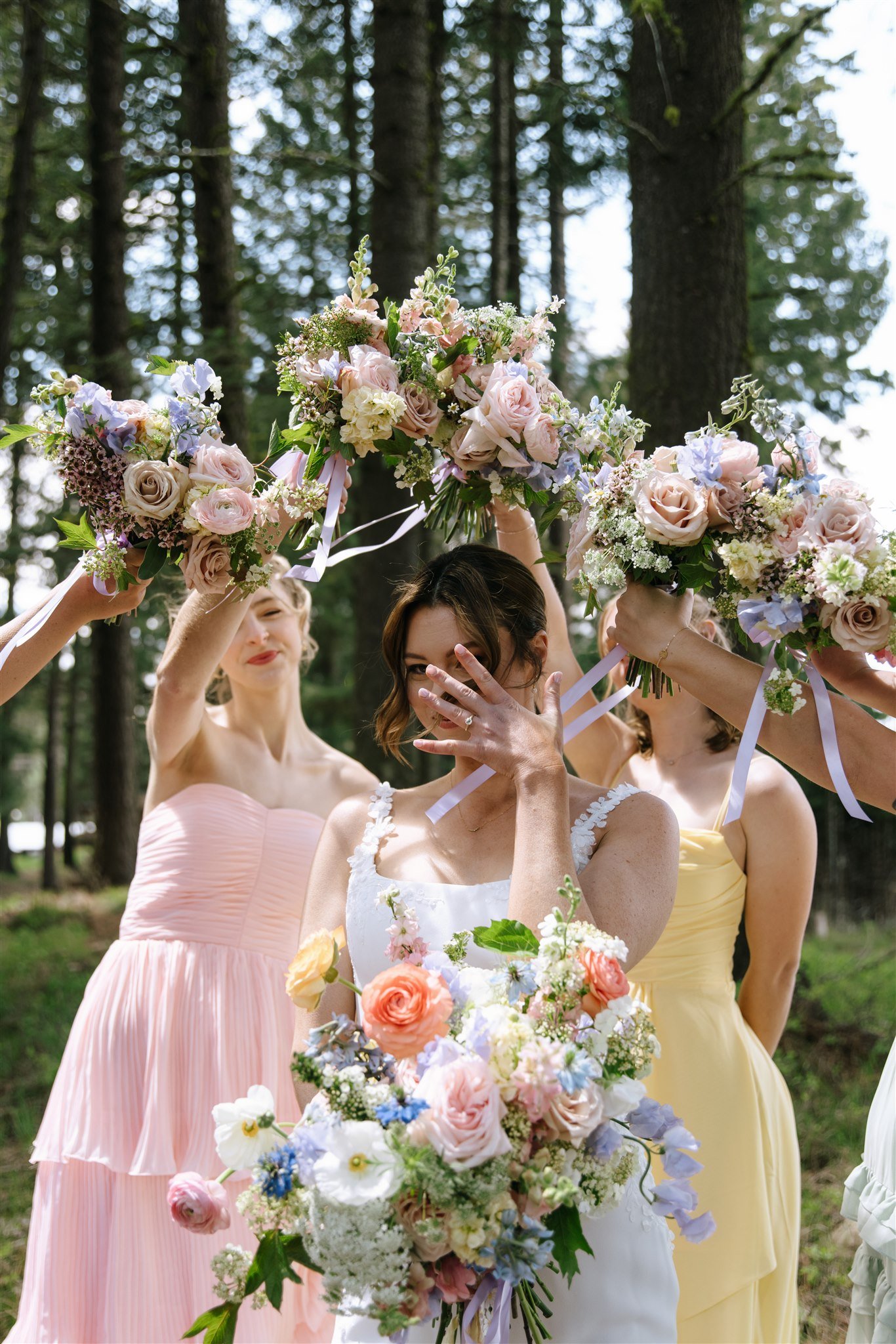 Women in pastel dresses holding bouquets of flowers in an outdoor woodland setting during a celebration.