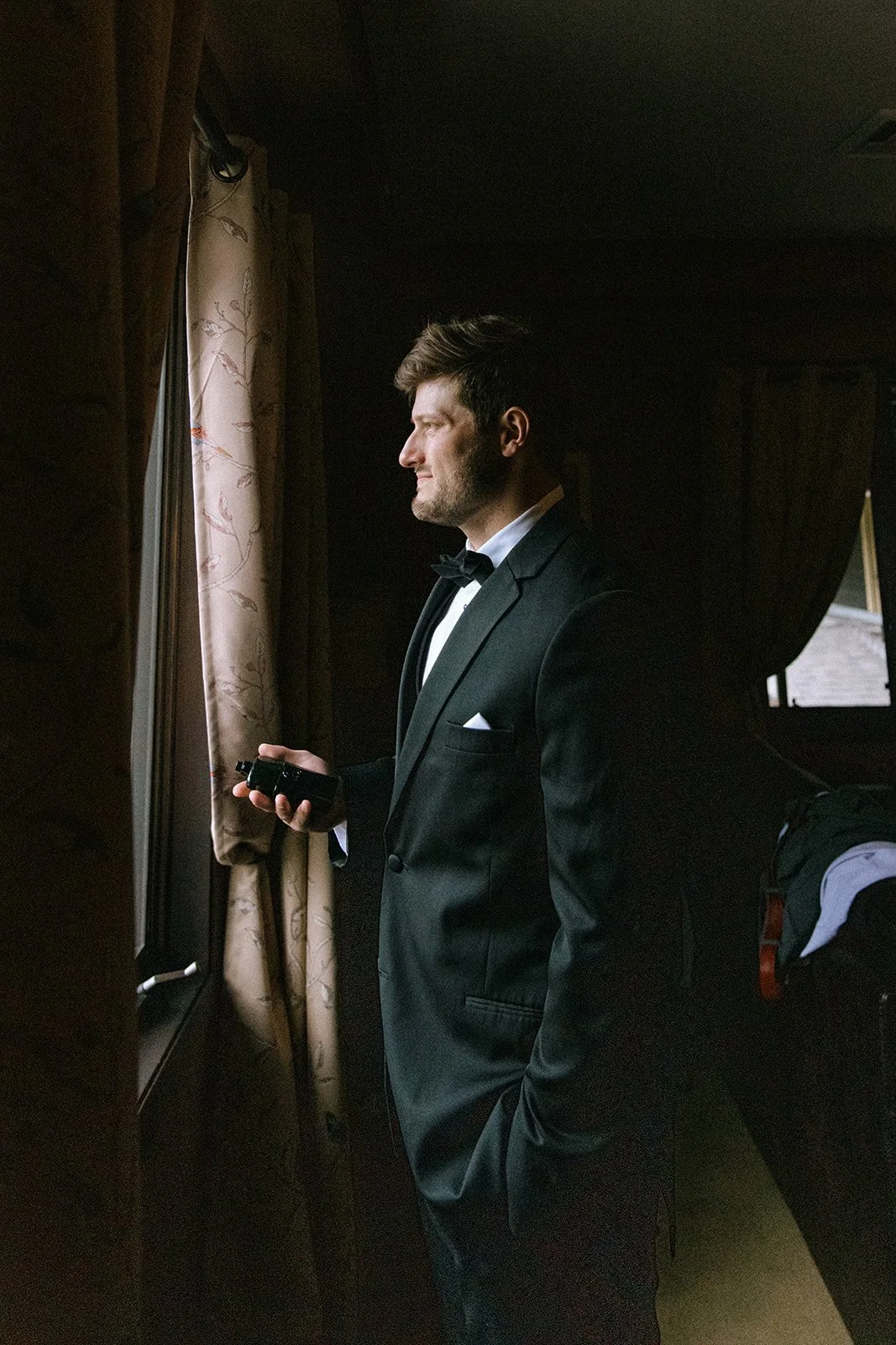 A man dressed in a black tuxedo with a white shirt and black bow tie, standing by a window with floral curtains, looking outside while holding a small object in his hand.