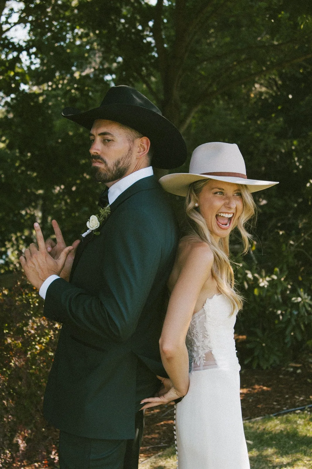 A man and woman standing back-to-back outdoors. The man is wearing a black cowboy hat and a black tuxedo, looking serious. The woman is wearing a white dress and a white wide-brim hat, laughing with her mouth open. They are positioned against a backg