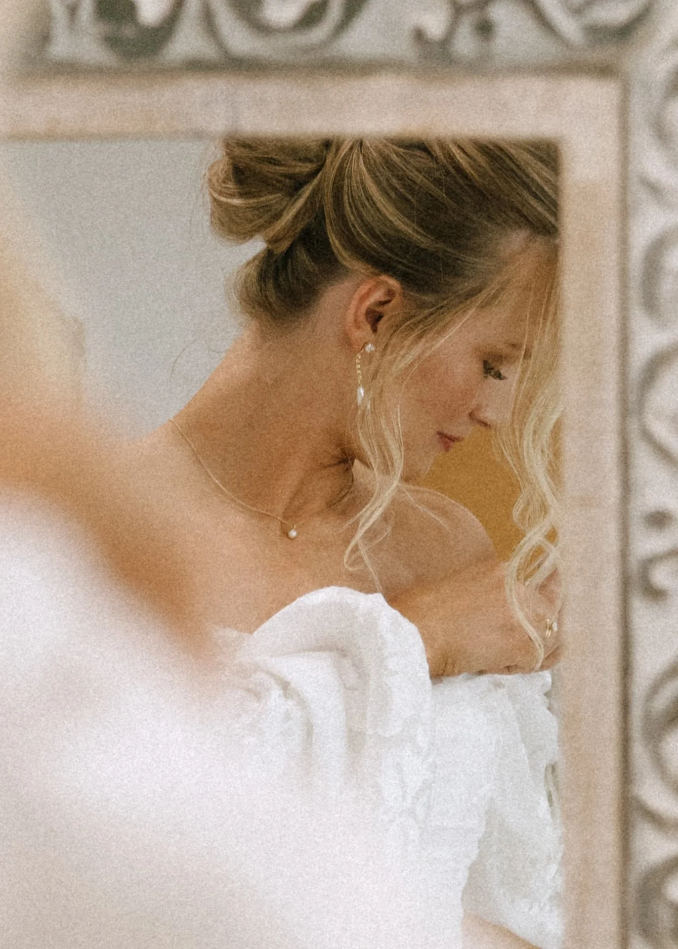 A woman with styled blonde hair in an updo and loose curls, looking downward, wearing earrings, a necklace, and a white dress with ruffles, standing in front of a mirror with decorative frame.