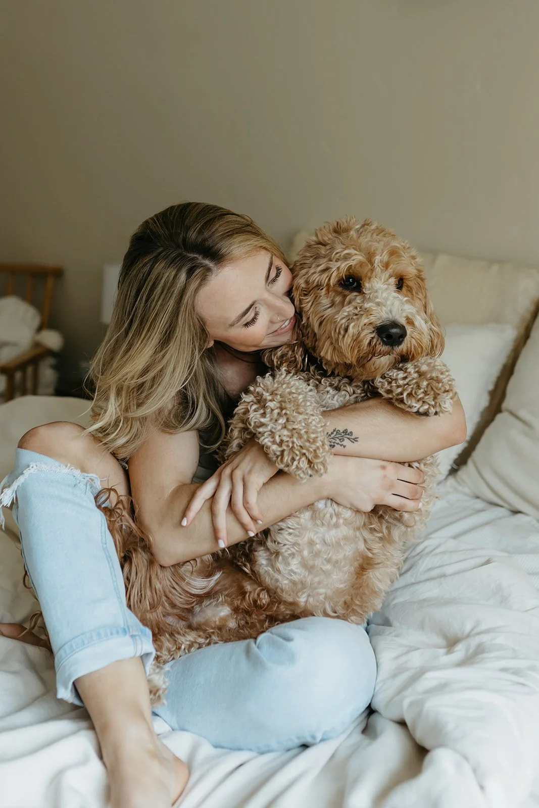 A woman hugging a curly-haired, tan and white dog on a bed in a cozy bedroom.