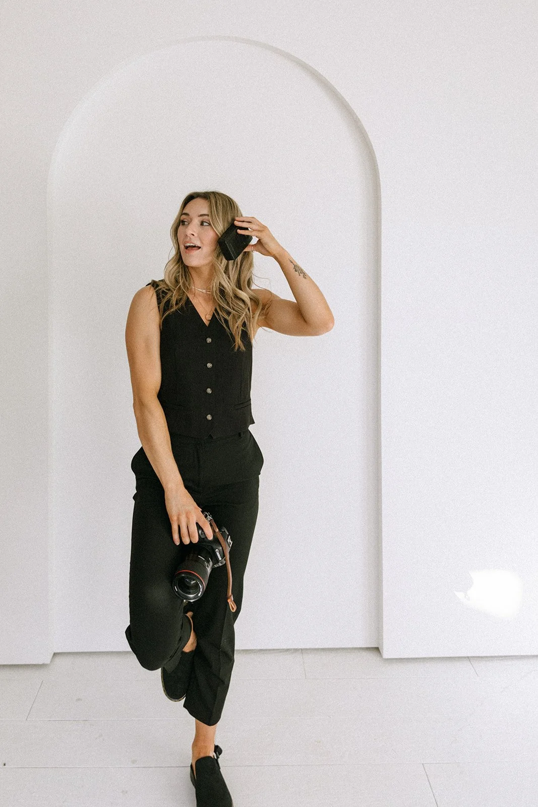 A woman with long blonde hair is standing against a white wall with an arch and talking on a cell phone. She holds a camera in her right hand and is dressed in a black sleeveless top and black pants.
