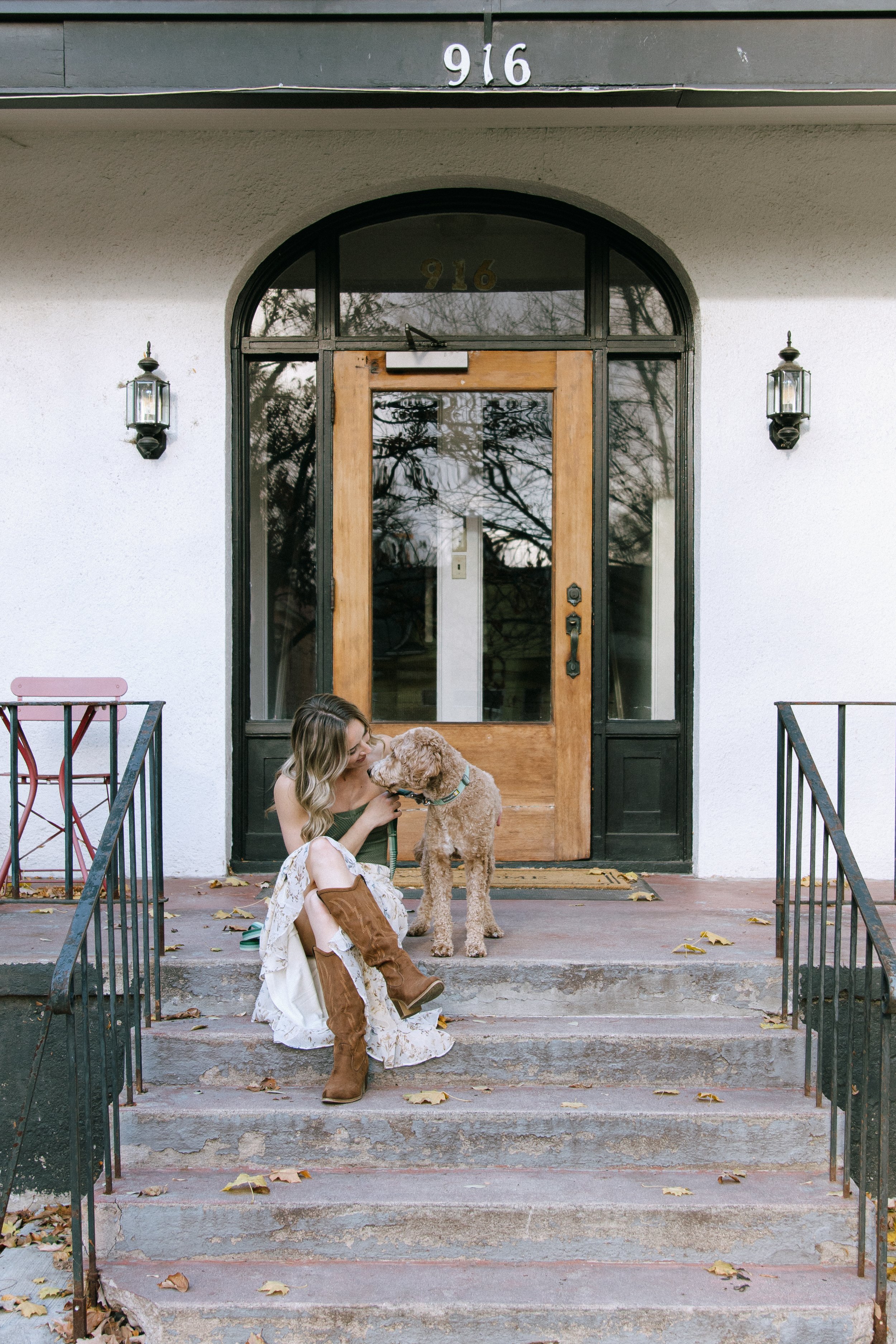 A woman sitting on concrete stairs in front of a house, kissing a large curly-haired dog. The house has a large wooden front door with glass panels and the number 916 above it. There are two outdoor lanterns on either side of the door. Fall leaves are scattered on the stairs.
