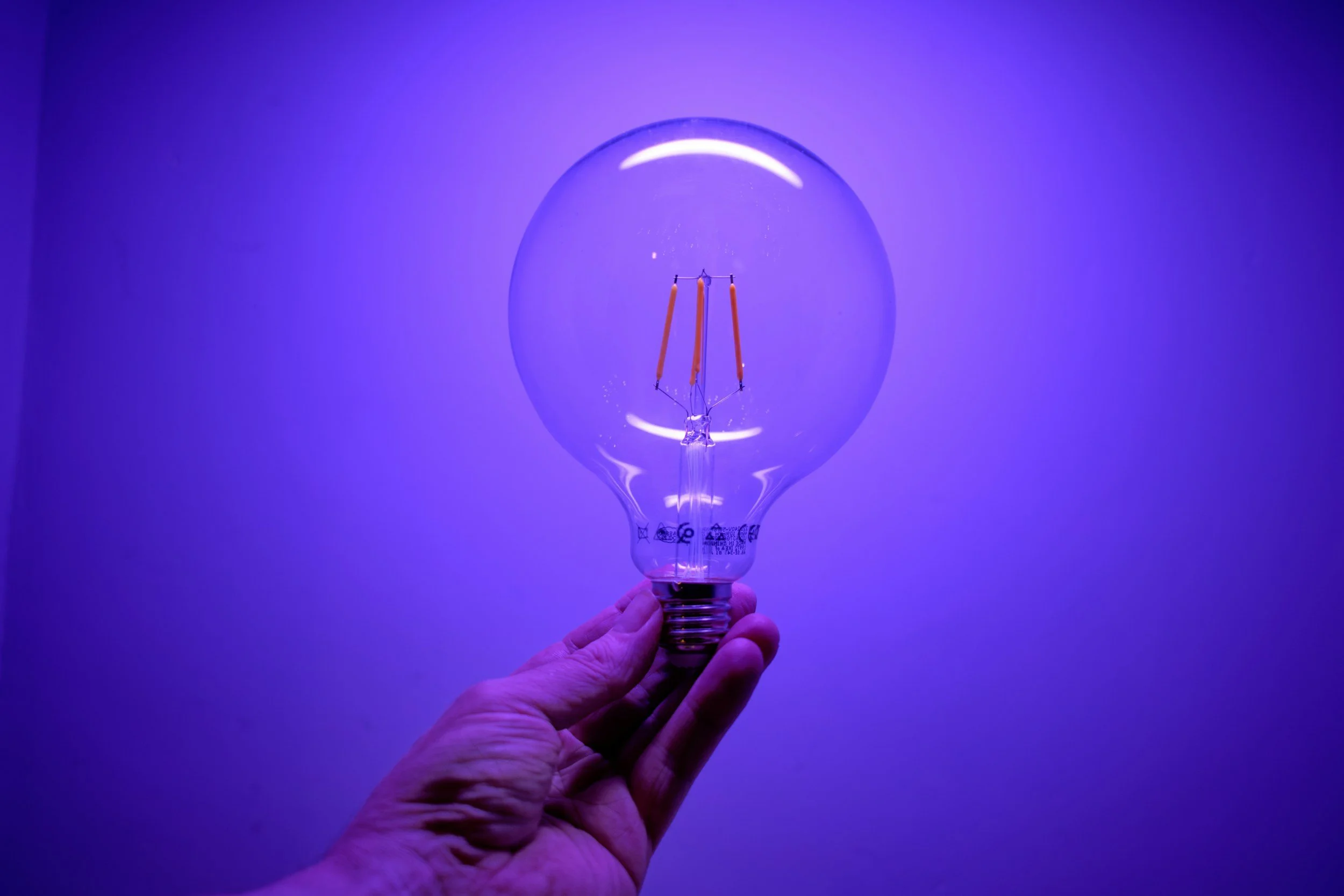 Hand hält eine Glühbirne mit sichtbarem Glühfaden gegen einen bläulichen Hintergrund.