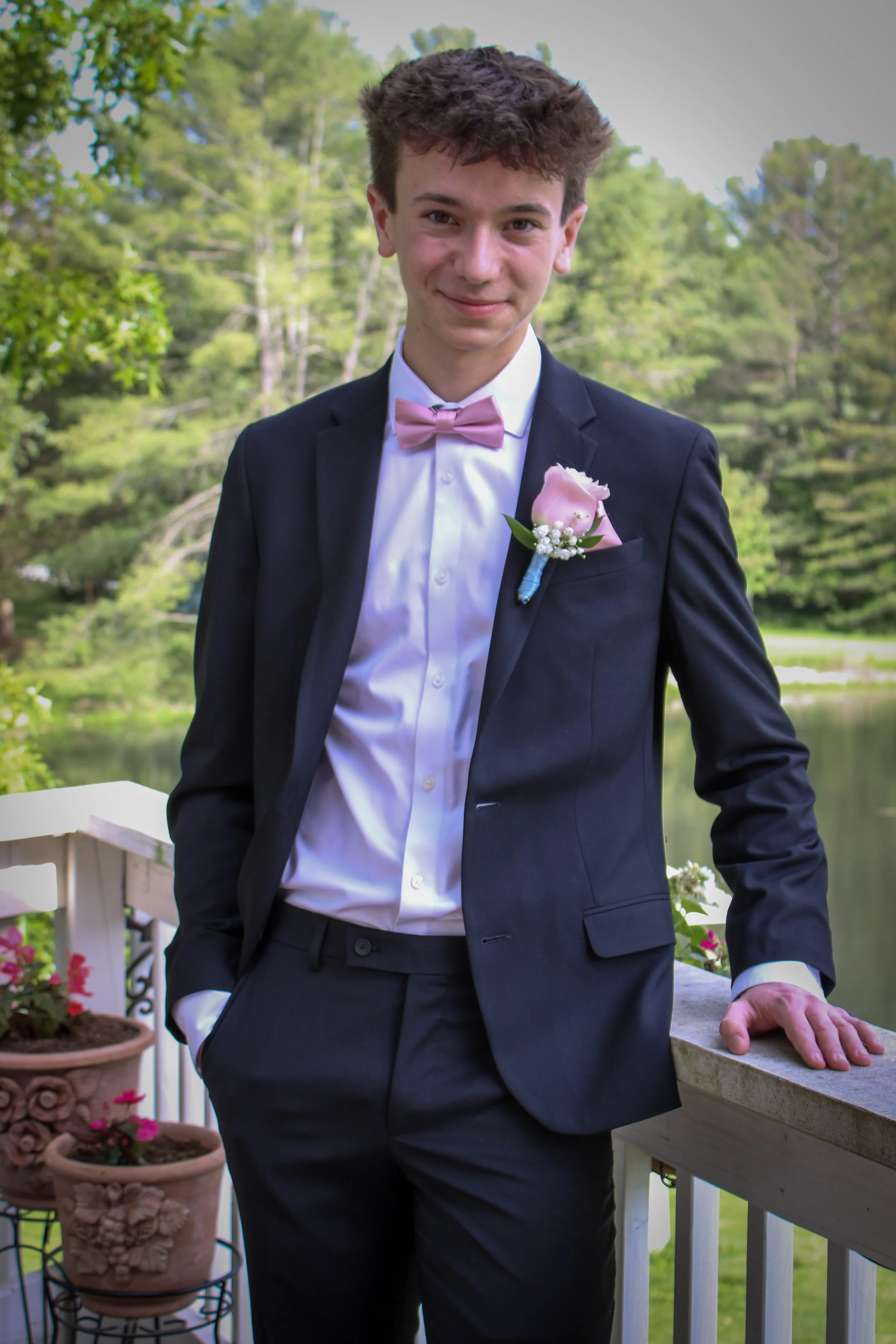 Founder Everett Sherfick in a tuxedo with a pink bow tie and boutonnière, standing on a deck outdoors near a body of water, surrounded by trees and potted flowers.