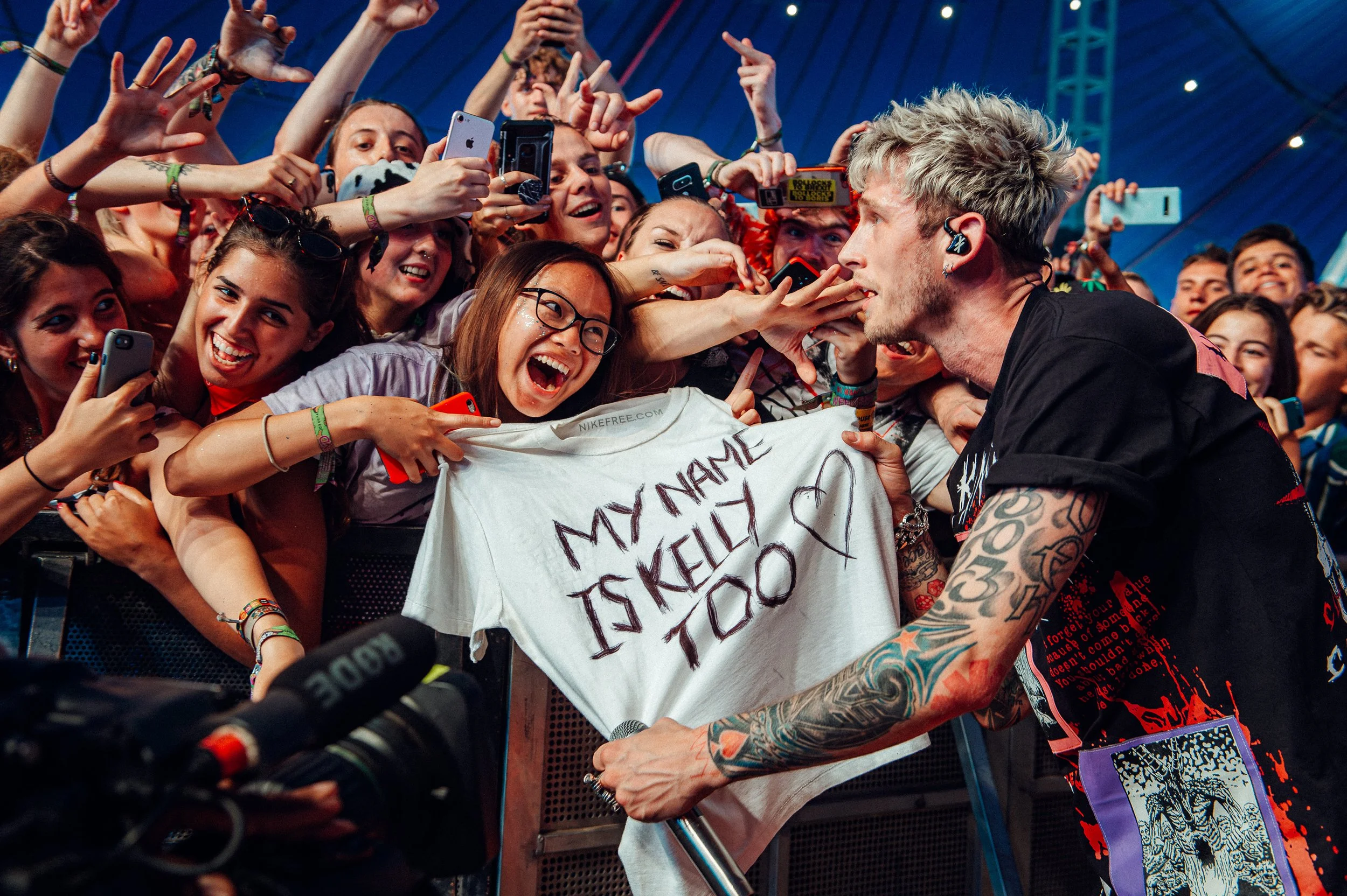 Machine Gun Kelly  interacting with a crowd of excited fans at a concert, holding a sign that says 'My name is Kelly To' amid a sea of people taking pictures and reaching out.