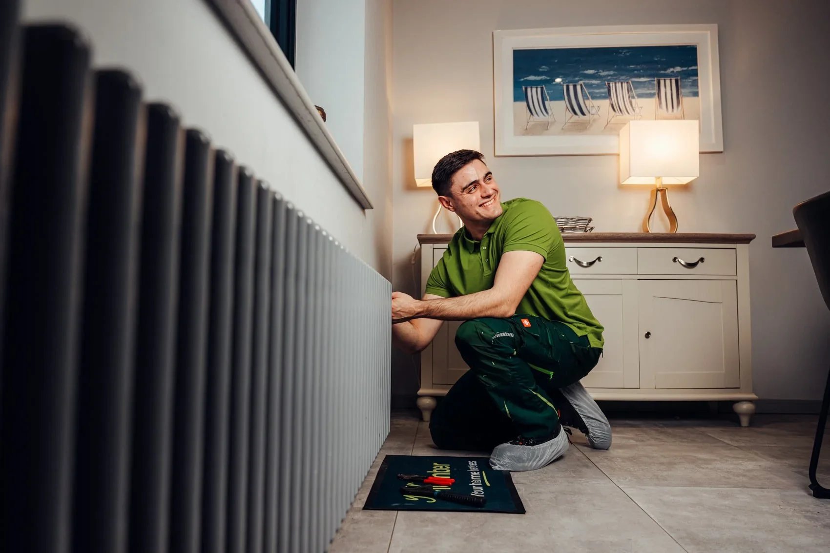 A man in a green polo shirt and work pants kneeling on the floor, working on a radiator in a room with a sideboard and framed beach scene on the wall.