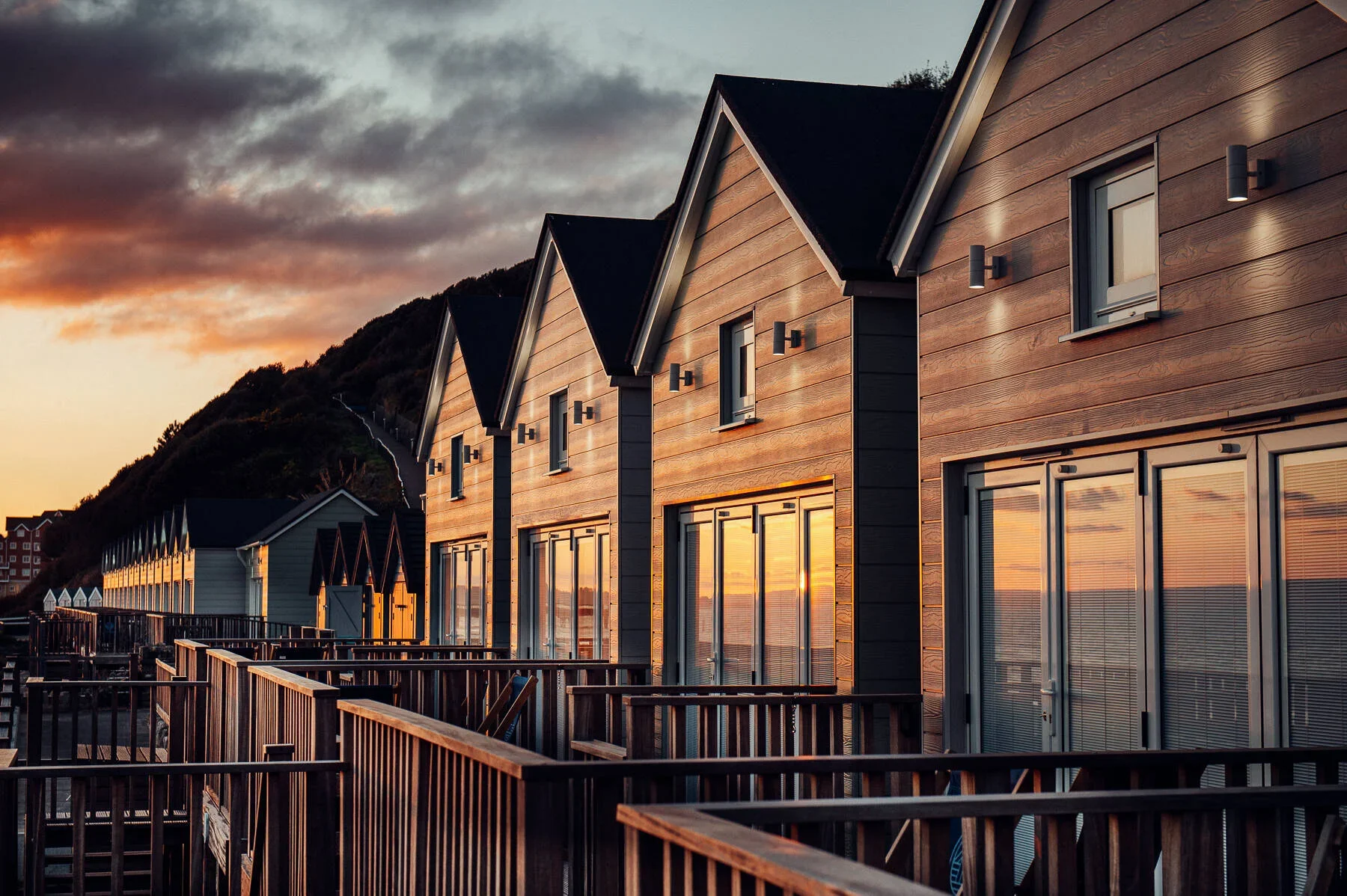 Lifestyle Photography - Beach huts at Boscombe. Row of modern wooden houses with large glass windows reflecting the sunset, shoulder mountain in the background, at sunset.