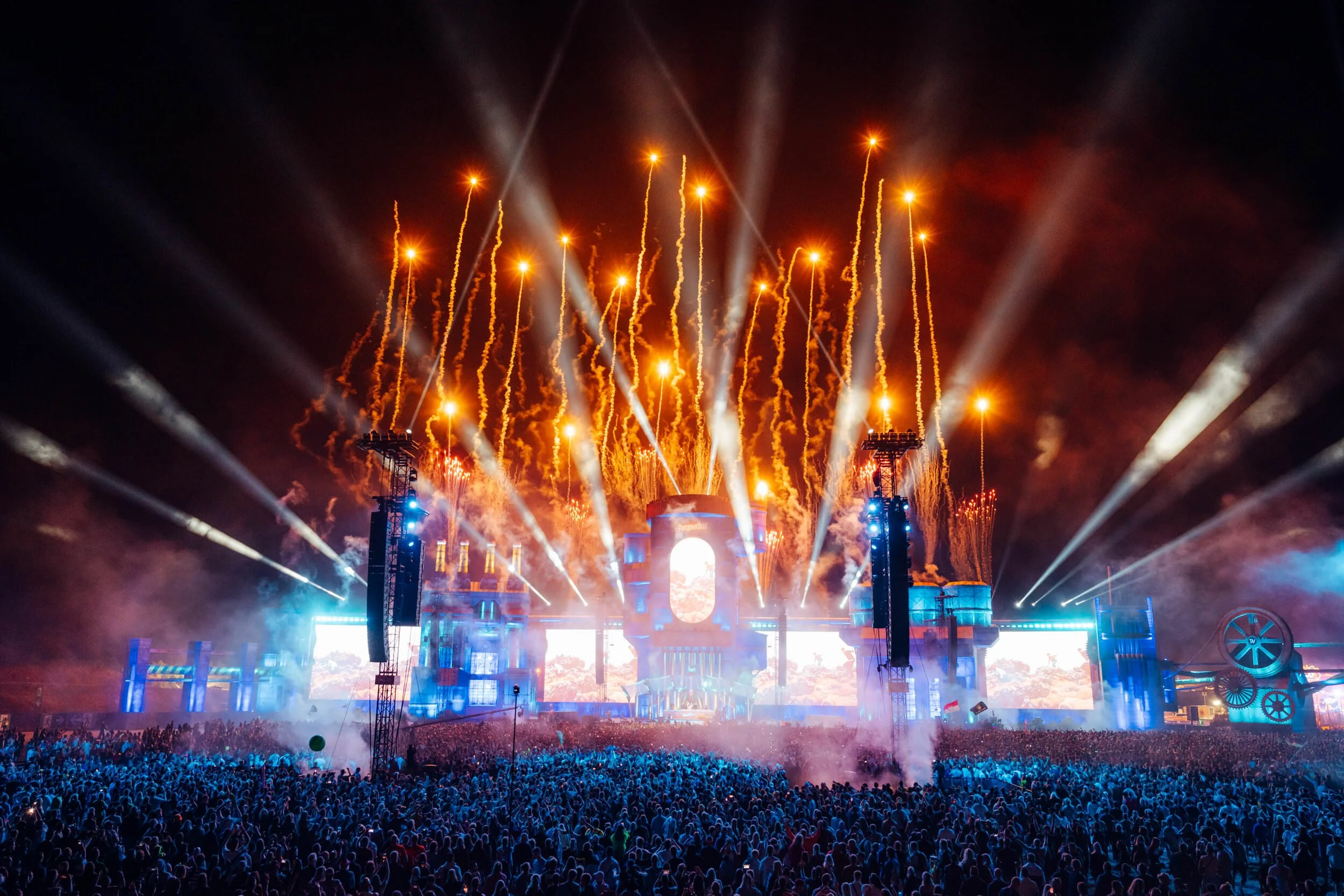 Parookaville - Steve Aoki Set shows people watching a fireworks display at night, with bright orange and white fireworks illuminating the sky and stage lighting creating a vibrant atmosphere.