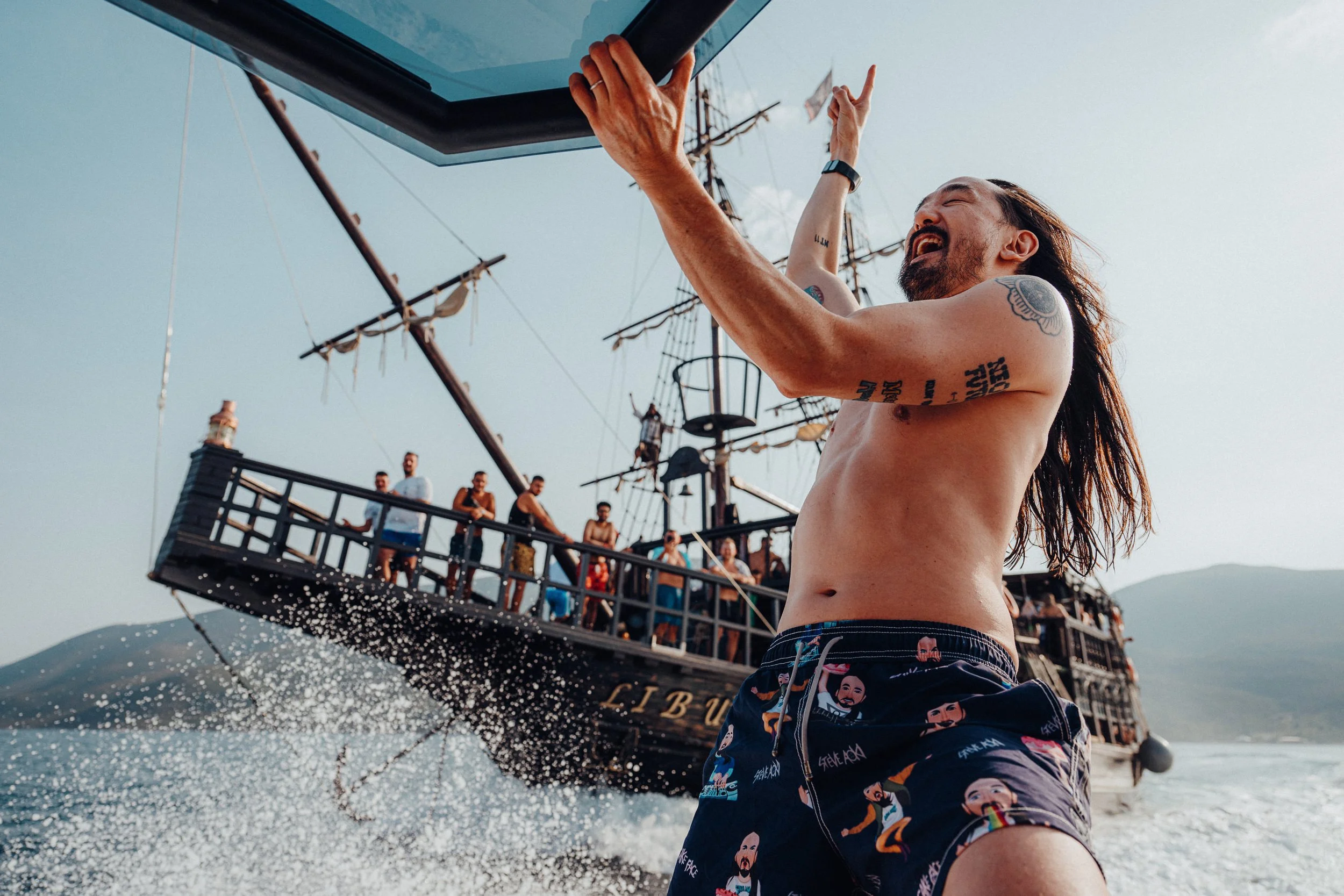Steve Aoki enjoying time on the water, with a historic sailing ship and a group of people in the background taken in Albania 2024