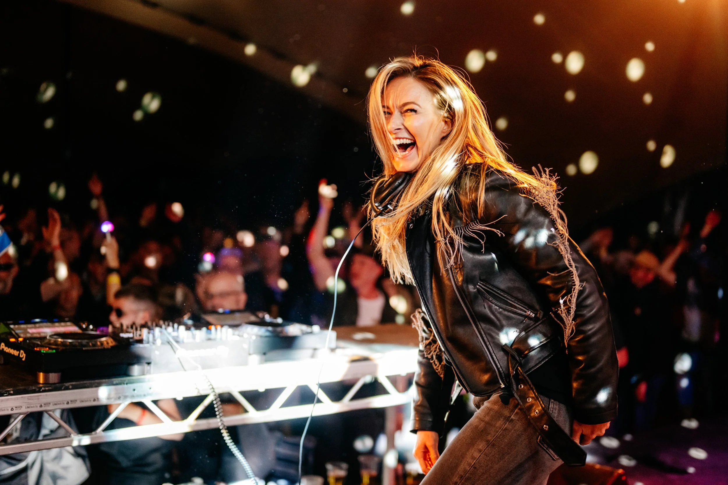 DJ with blonde hair wearing a black leather jacket, standing at a mixing console, smiling and singing into a microphone, party crowd in the background with people dancing and raised hands.
