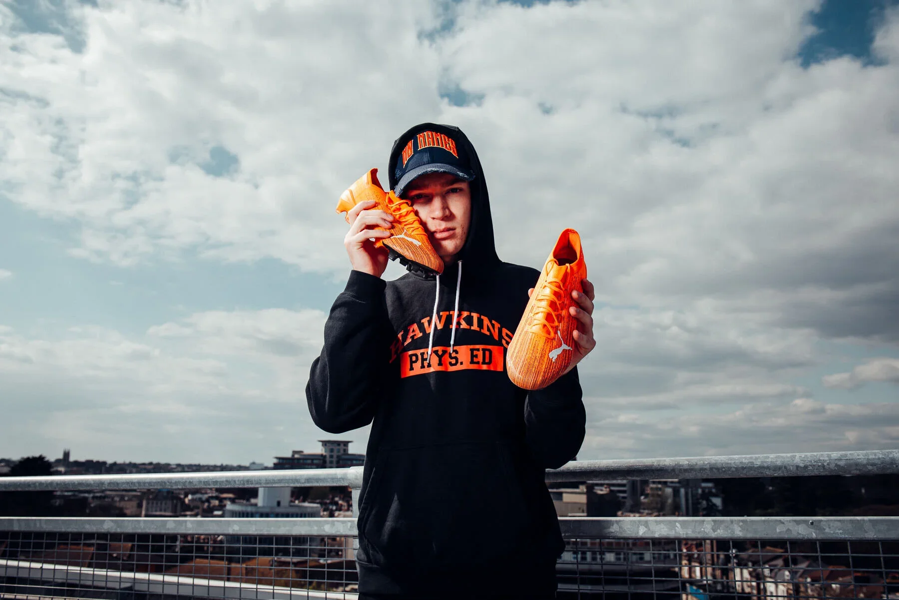 A young man in a black hoodie and cap holding a pair of orange Puma sneakers on an outdoor rooftop.