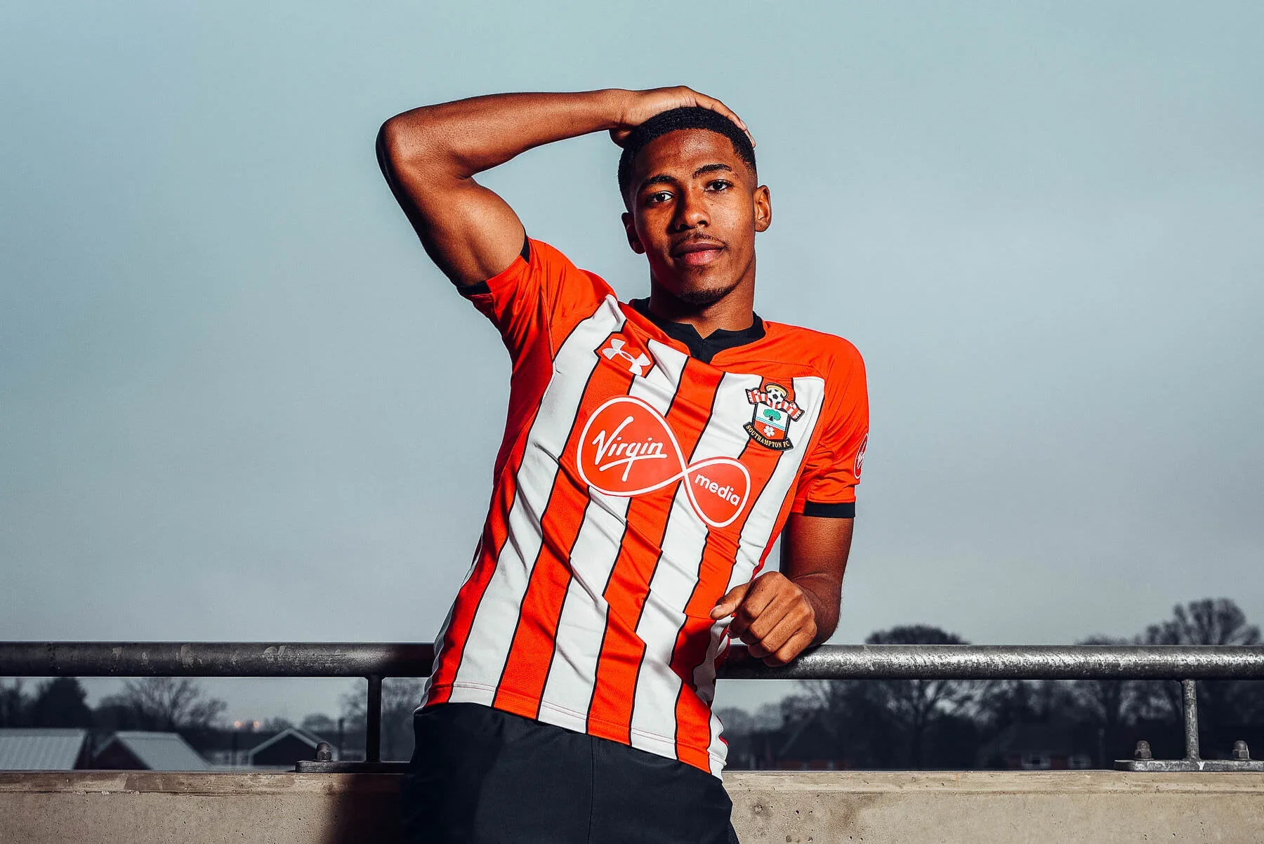 Young male soccer player in a red and white striped jersey posing with hand behind his head and the other resting on a railing, outdoors on a cloudy day.