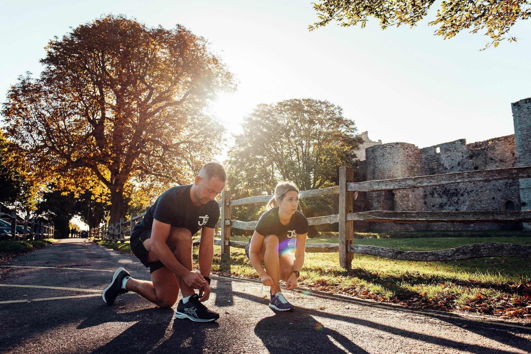 Fitness PT Brand shoot by James Bridle Photography UK. A man and a woman in athletic clothing tying their shoelaces outside during sunrise or sunset, with trees and an old stone castle in the background.