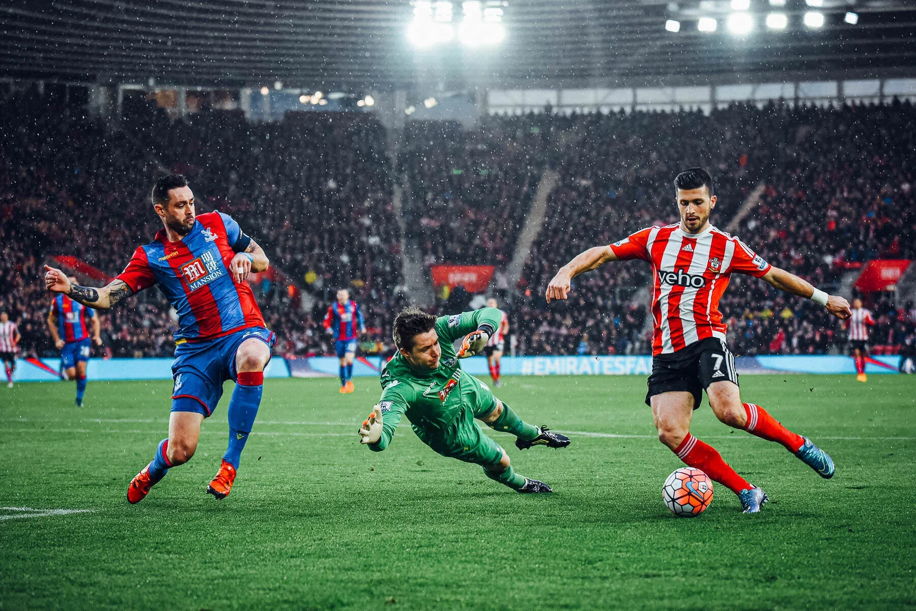 A soccer match with three players competing for the ball on a grass field, with a stadium full of spectators in the background under rainy weather.