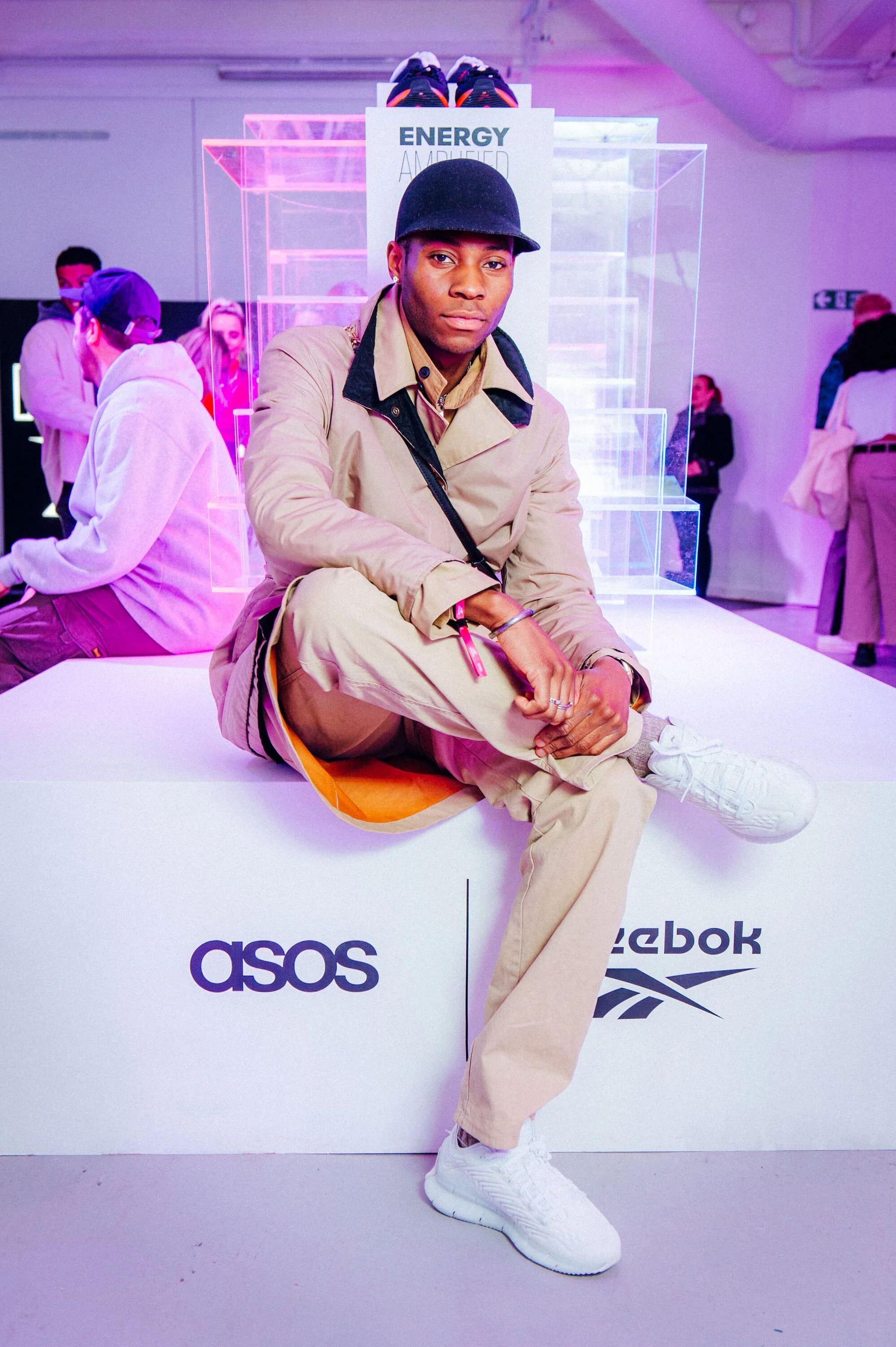 A person sitting on a white platform with the logos 'Asos' and 'Reebok', in an event space illuminated with colorful lighting. The person is wearing a black cap, beige jacket, beige pants, and white sneakers. Captured by James Bridle Photography 