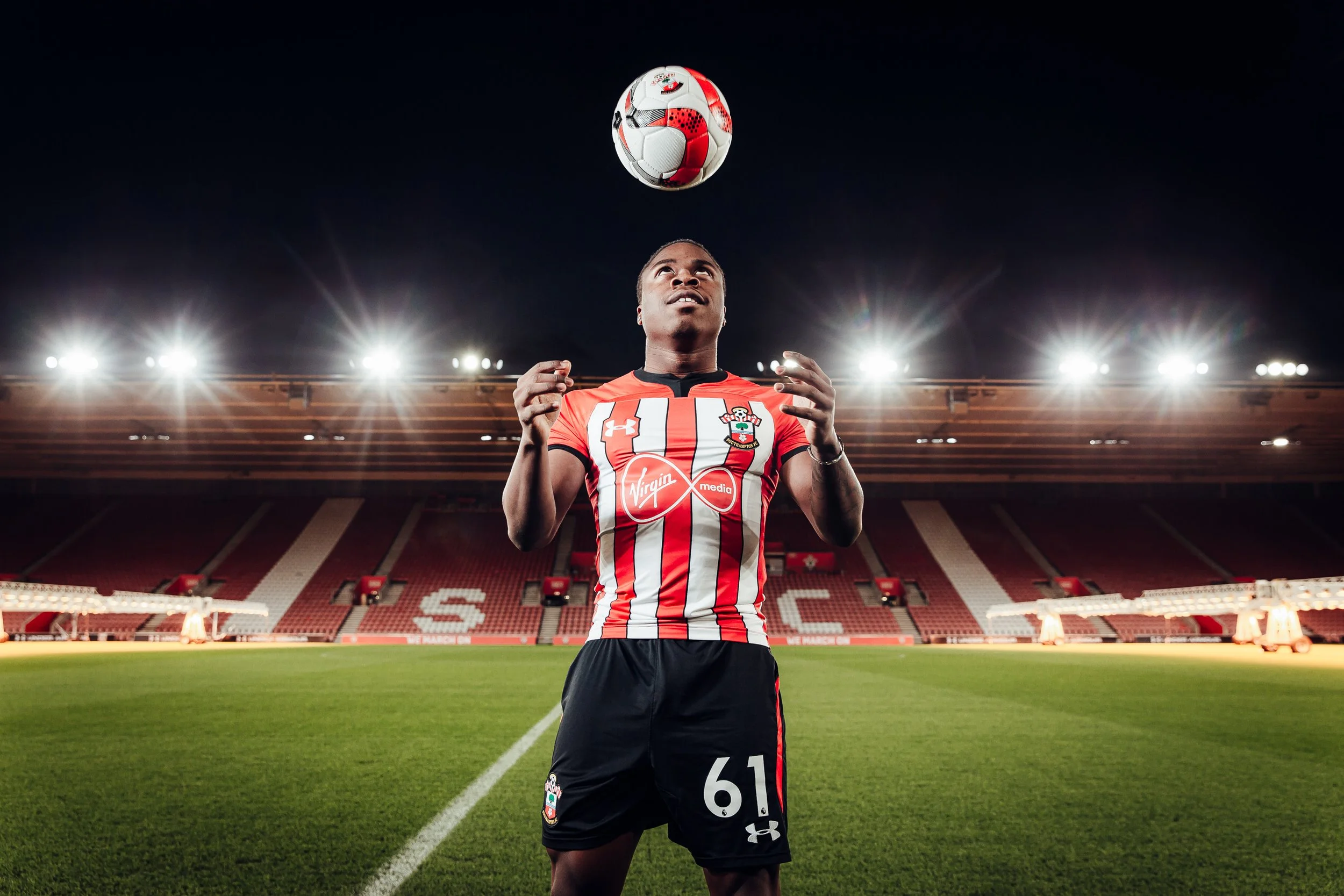 Michael Obafemi in a red and white striped Southampton FC jersey with the number 61, standing on the field in a stadium at night, preparing to catch a football. Captured by James Bridle during a player shoot at Southampton FC stadium, St Marys