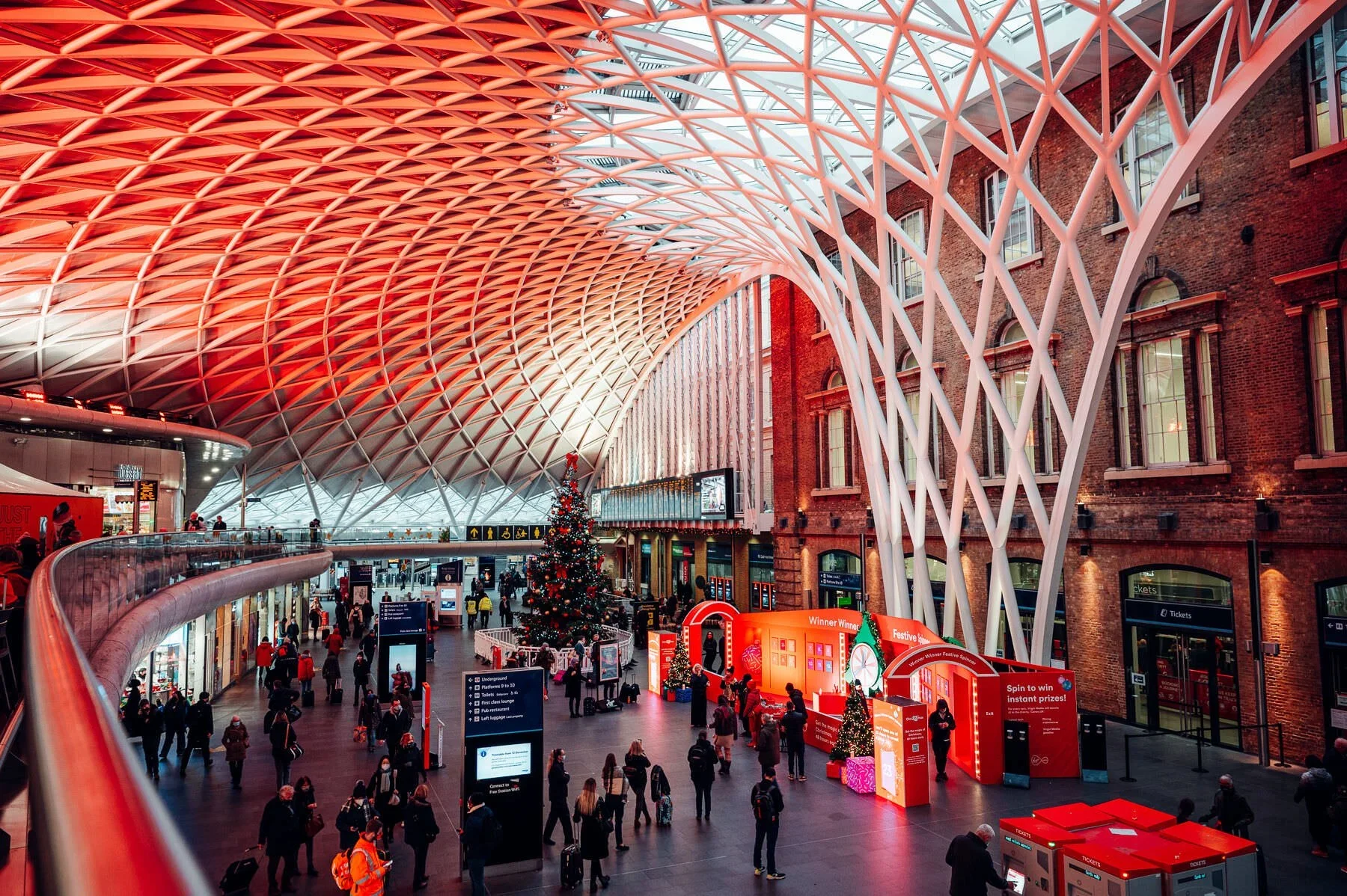 Interior of a Kings Cross Train station decorated for Christmas with a large Christmas tree, red and green decorations, and a modern architectural ceiling with geometric patterns. shot for Audience London, in association with Virgin Media.