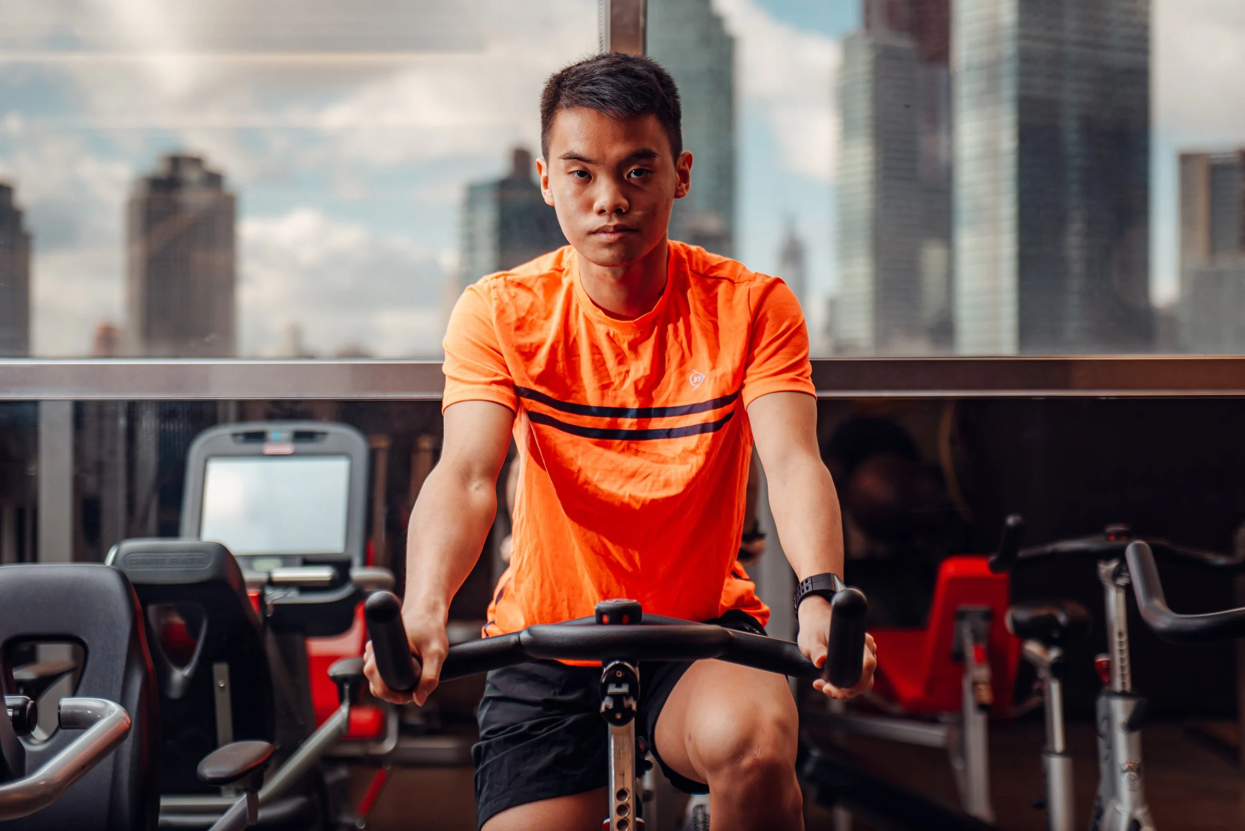Dunlop Commercial Shoot in NYC Long Island Captured by James Bridle. Eain Yow Ng in an orange t-shirt and black shorts riding a stationary exercise bike indoors with a cityscape view of tall buildings through large windows behind him.