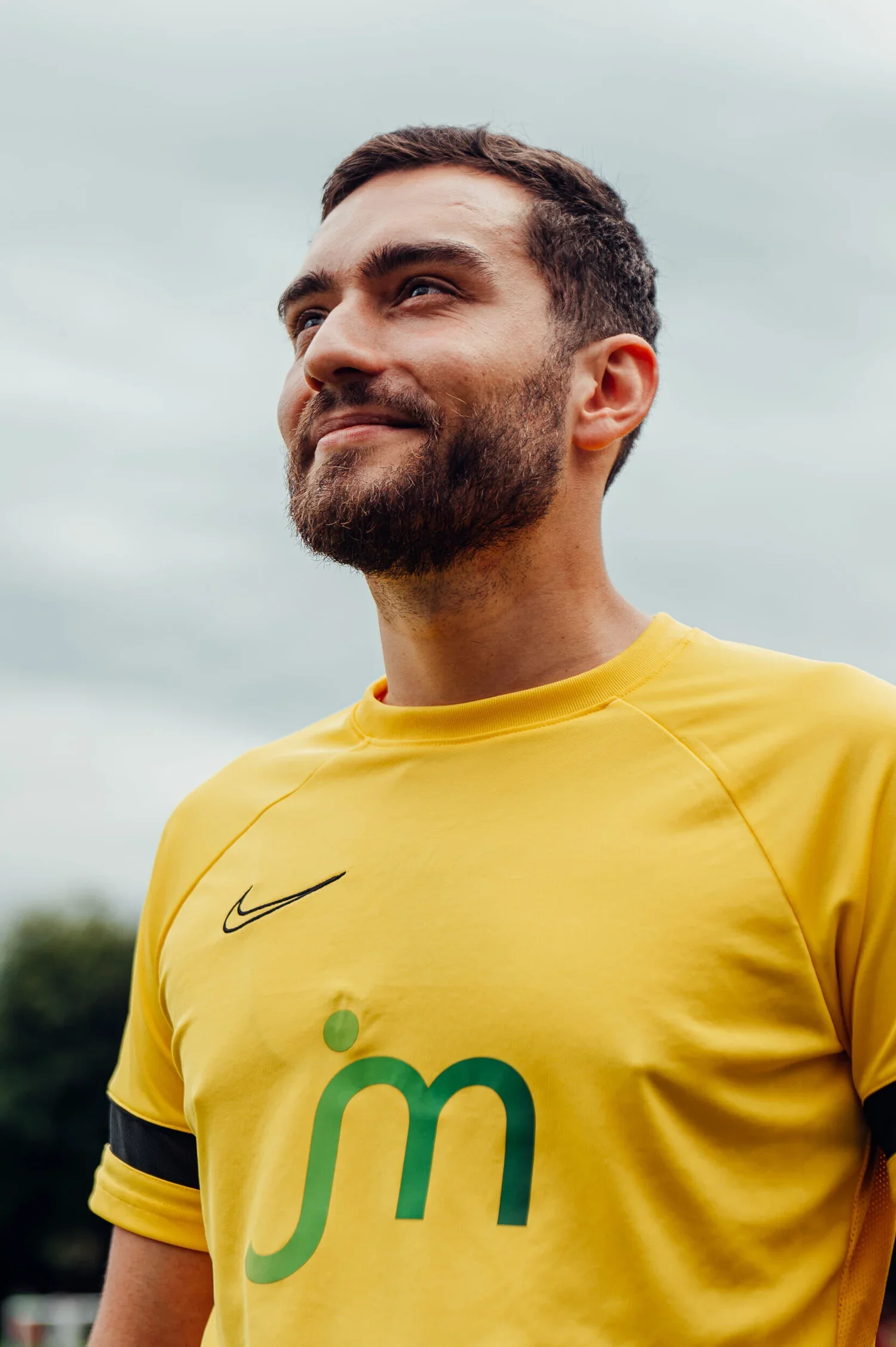 Nike Campaign by James Bridle Photography. A man with a beard wearing a yellow sports jersey, looking content outdoors with a cloudy sky in the background.
