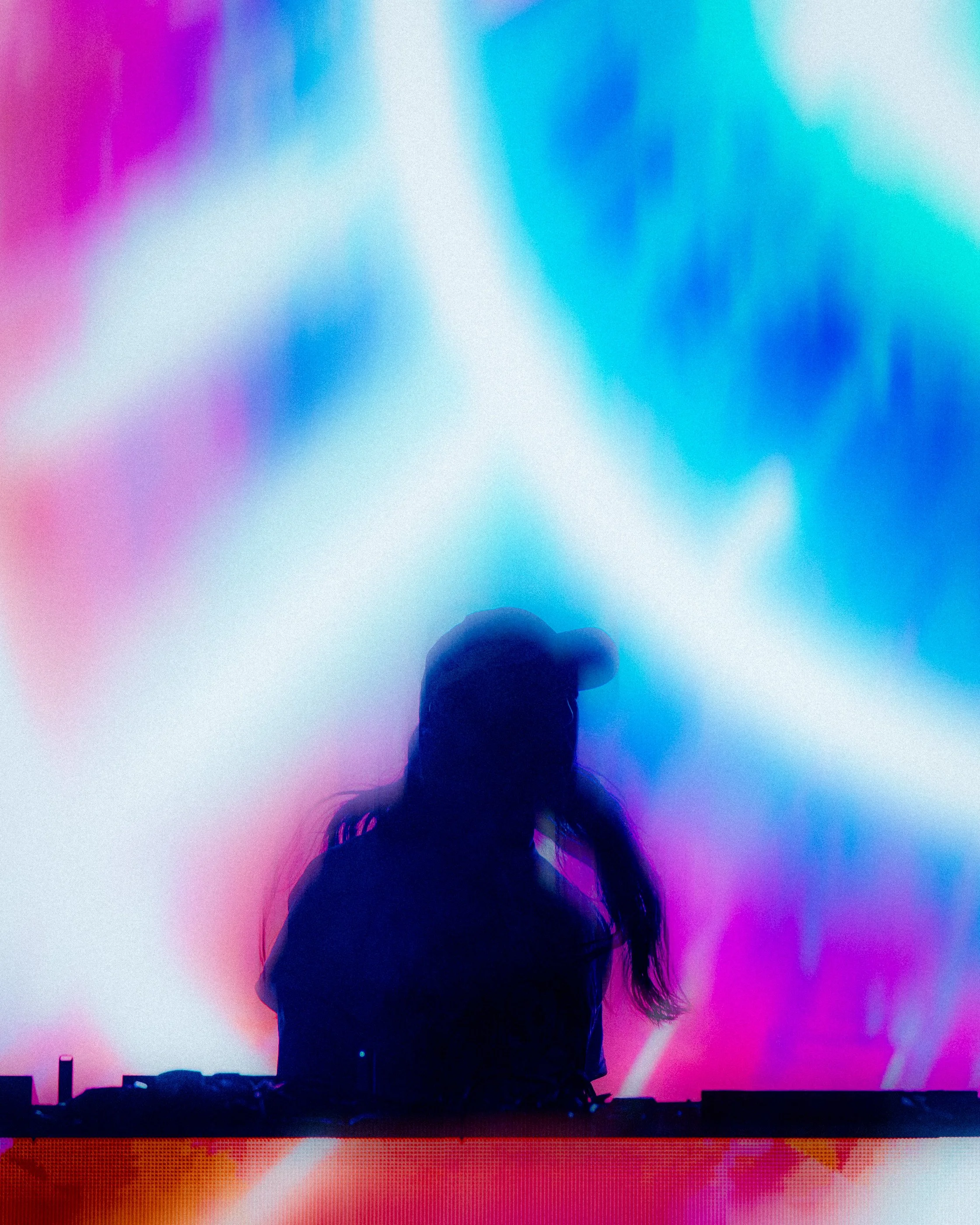 Steve Aoki Tour by James Bridle UK. Silhouette of Steve Aoki wearing a cap, standing behind DJ equipment against a vibrant, colorful background with pink, blue, and purple lights.