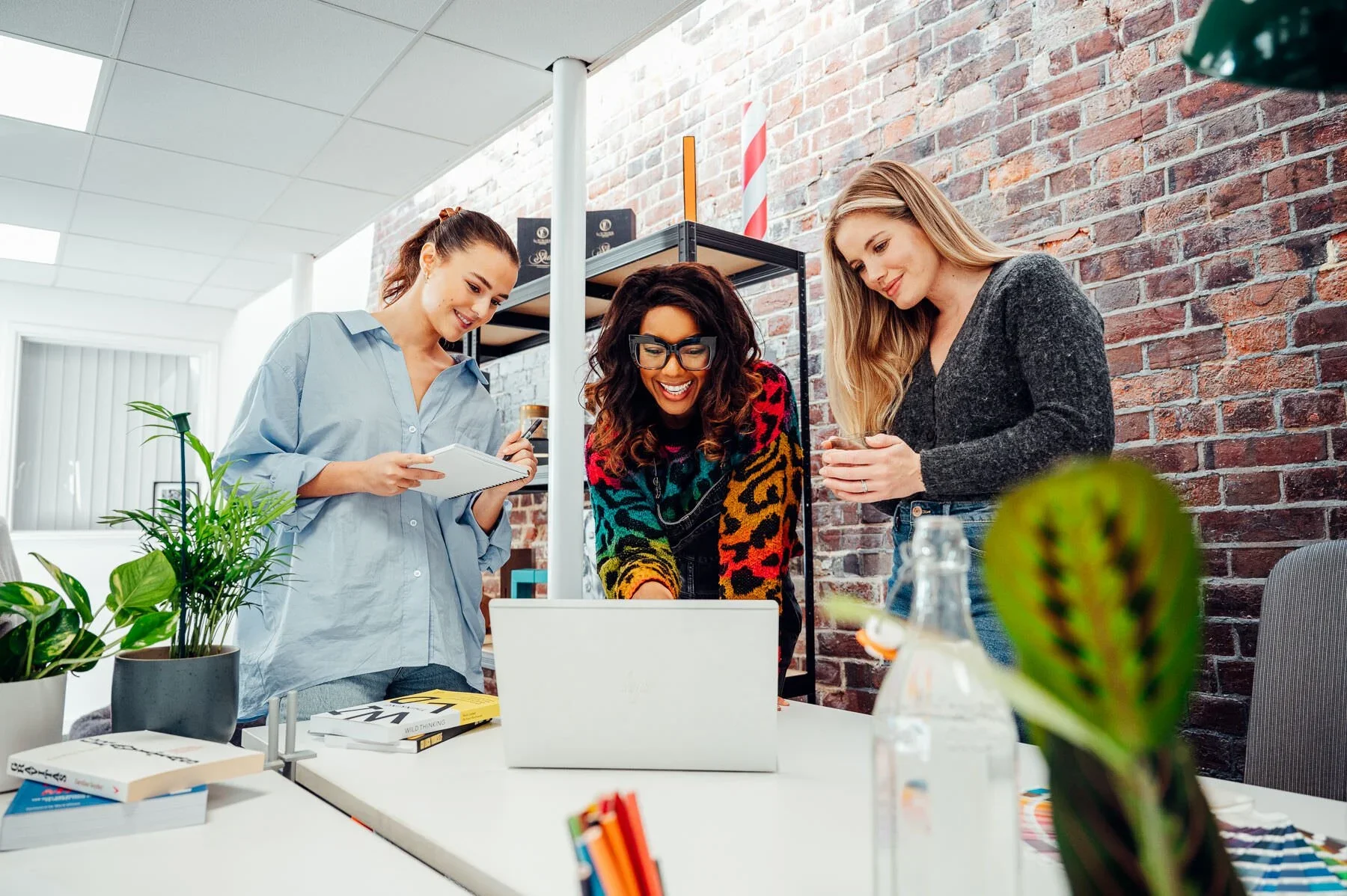 Three women are gathered around a laptop on a white table in a modern office with an exposed brick wall, discussing something with smiles and engagement. One woman is holding a notepad, another is looking at her phone, and the third is leaning over t