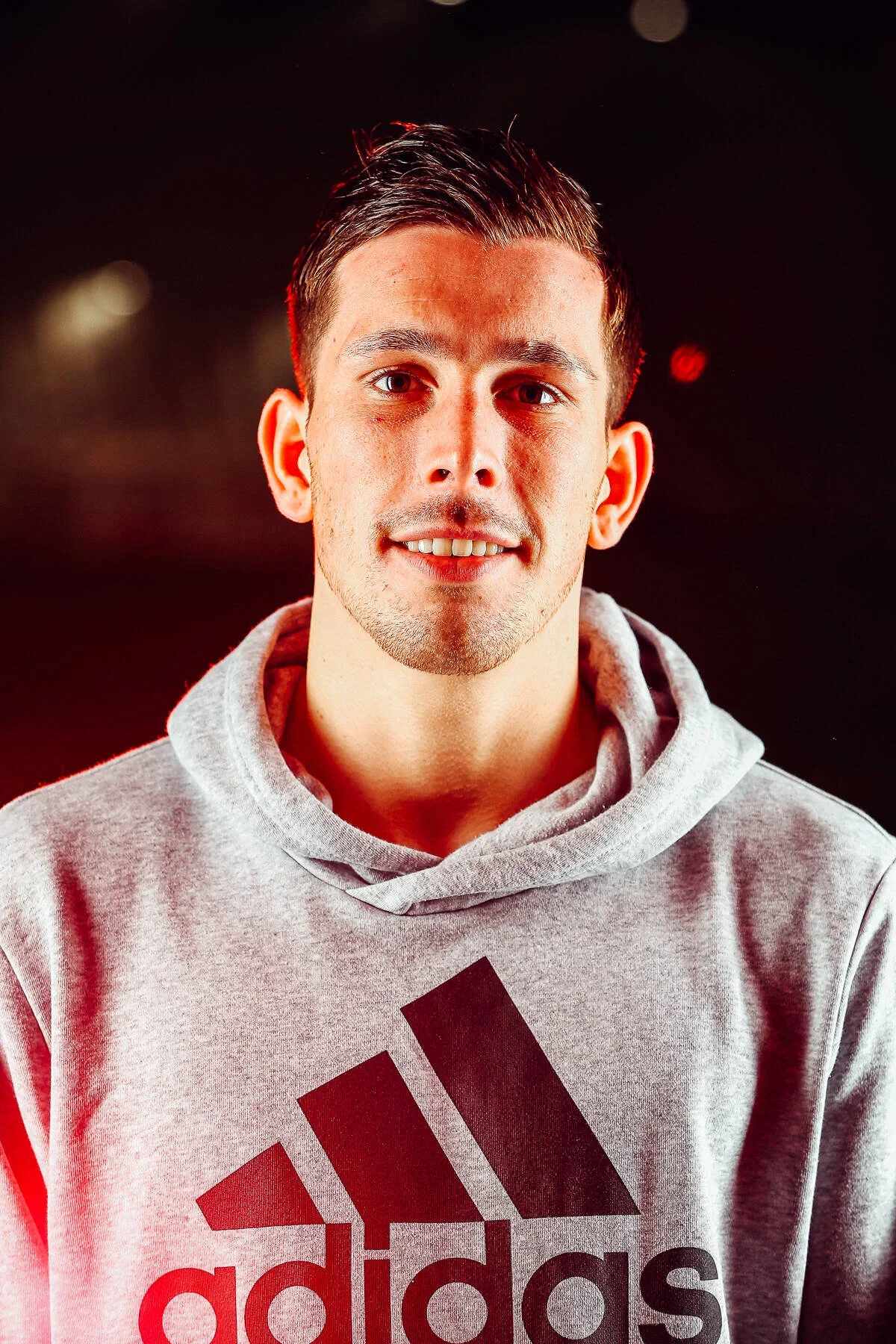 A young man with short dark hair, light skin, and a slight beard, wearing a gray Adidas hoodie, smiling at the camera against a dark background with some red and light bokeh.