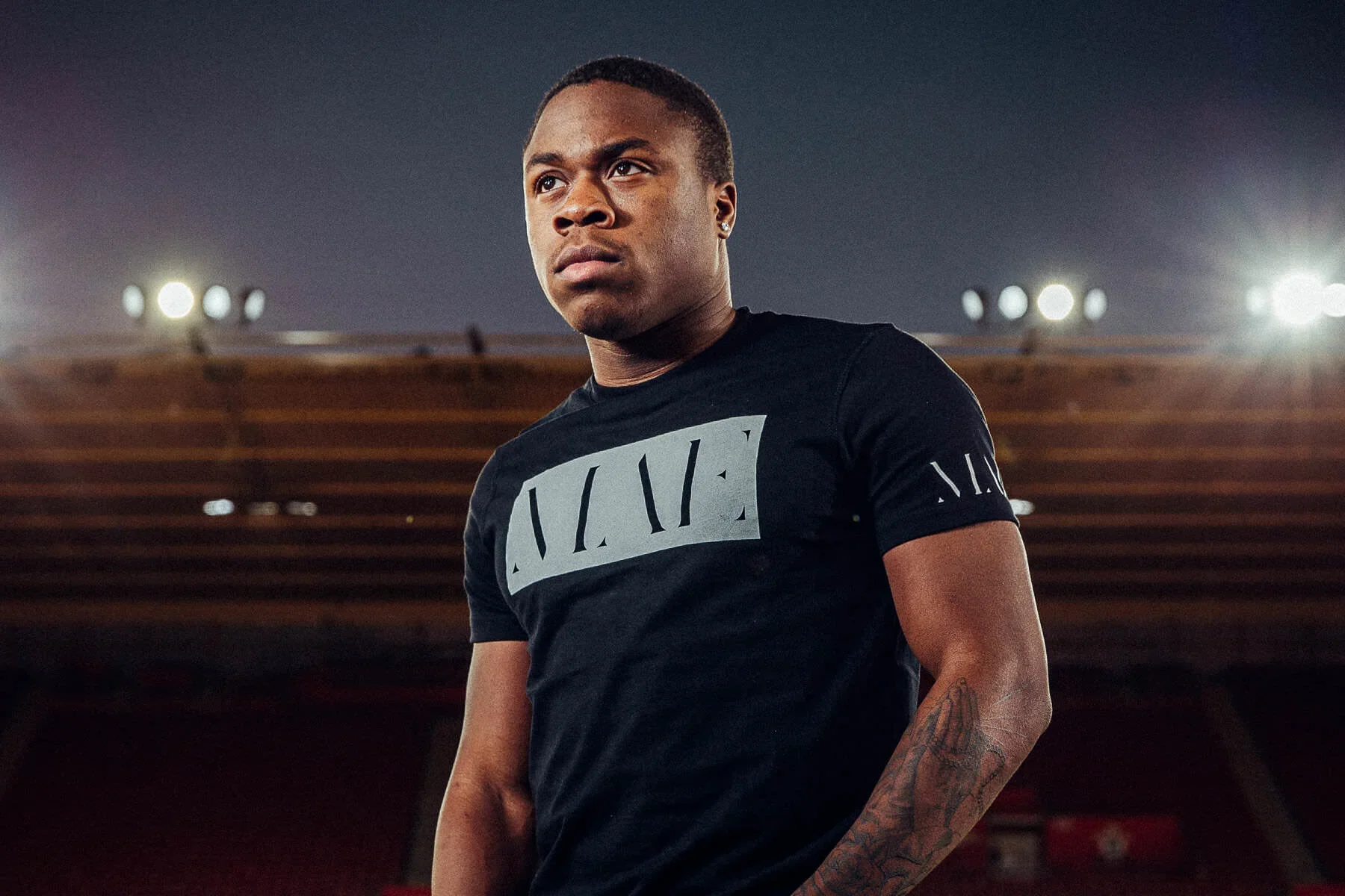 A young man with short hair and tattoos on his arm standing in a stadium at night, wearing a black T-shirt with a large graphic logo on the front.