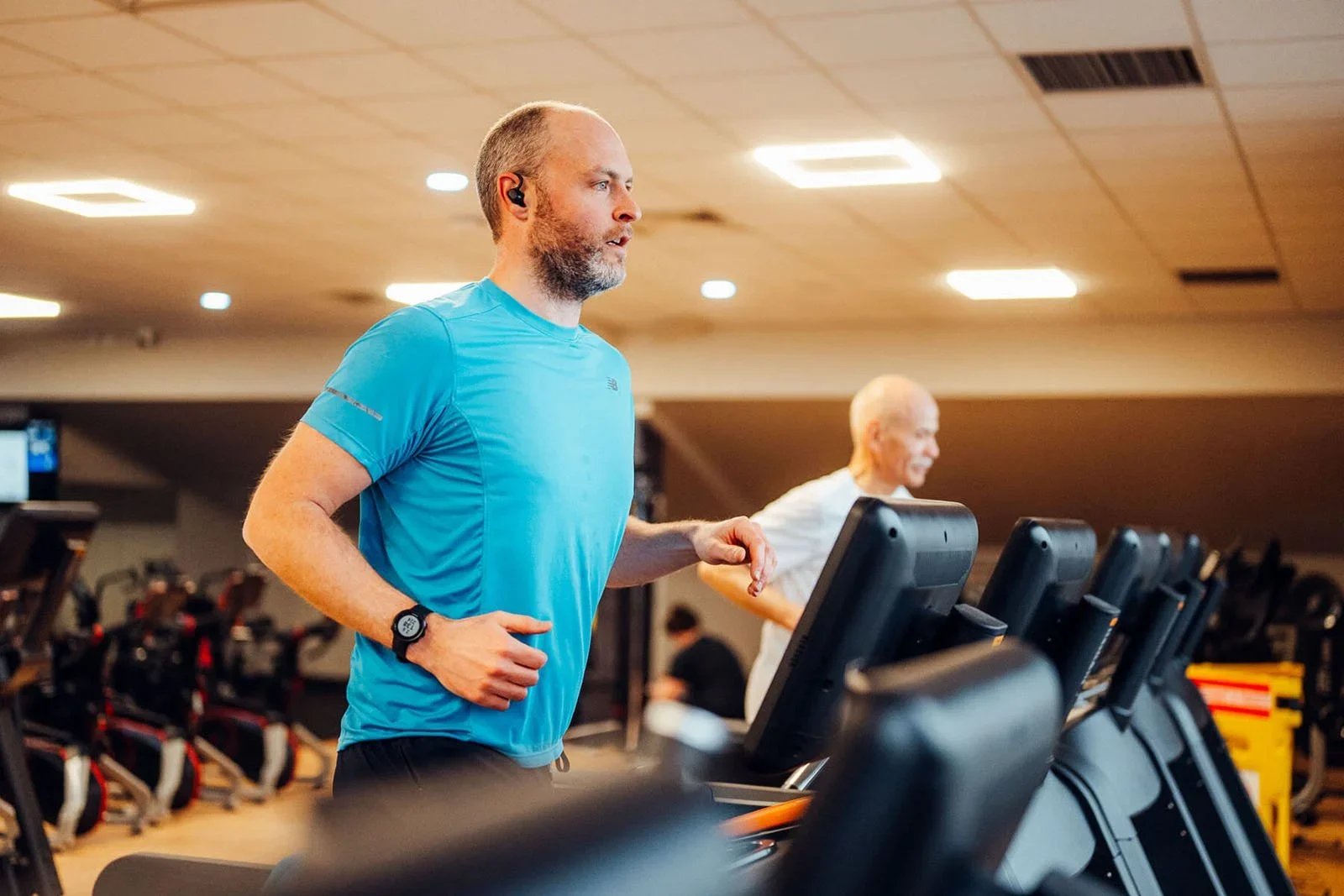 BHLive Fitness Campaign shot at a Local Centre by James Bridle Photography. Two men running on treadmills in a gym, one in a blue shirt and the other in a white shirt, with exercise equipment in the background.