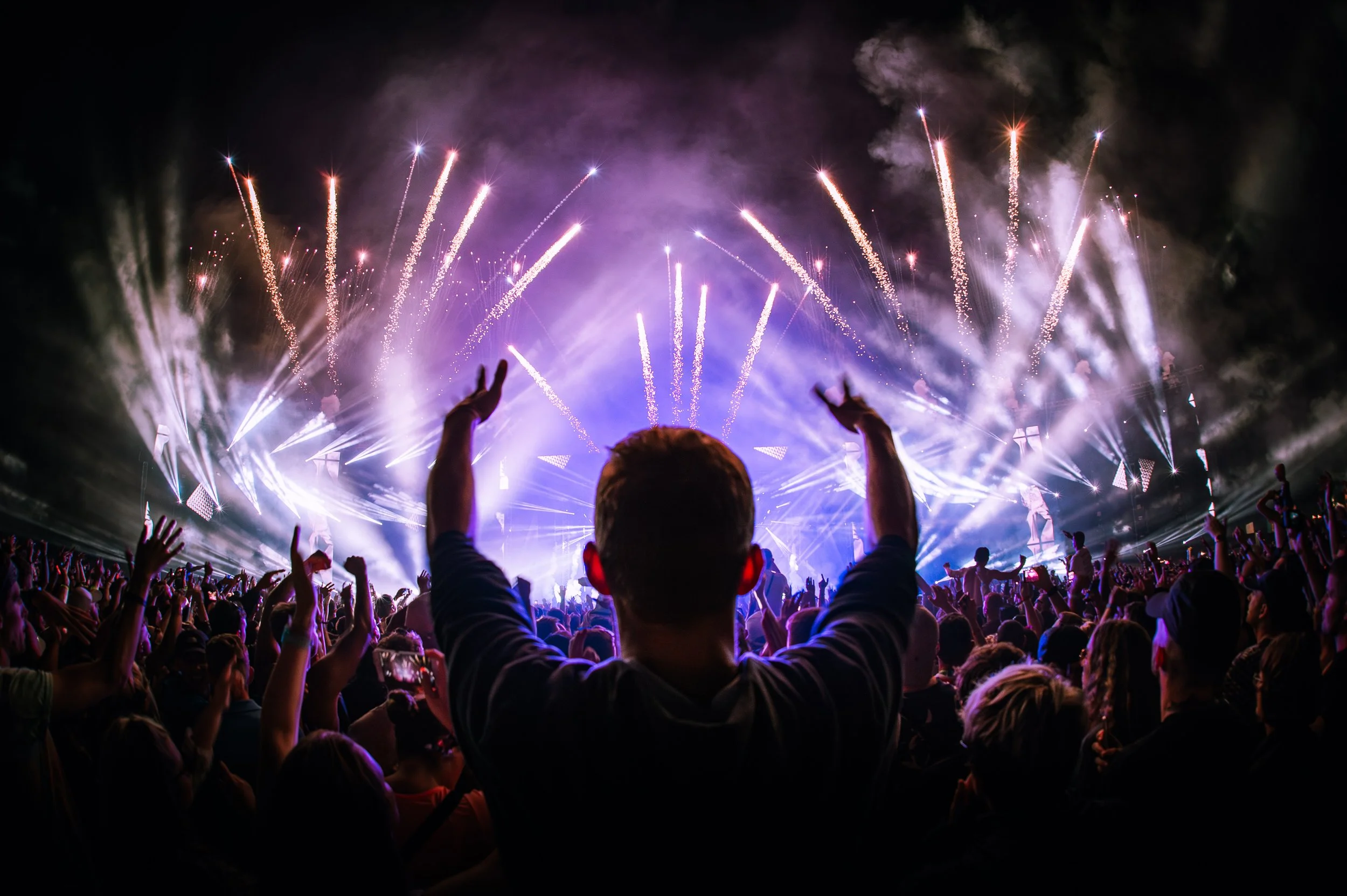 Creamfields 2017 captured by James Bridle Music Photographer showing a crowd at a concert or festival with fireworks exploding in the night sky, with a person in the foreground raising their hands.