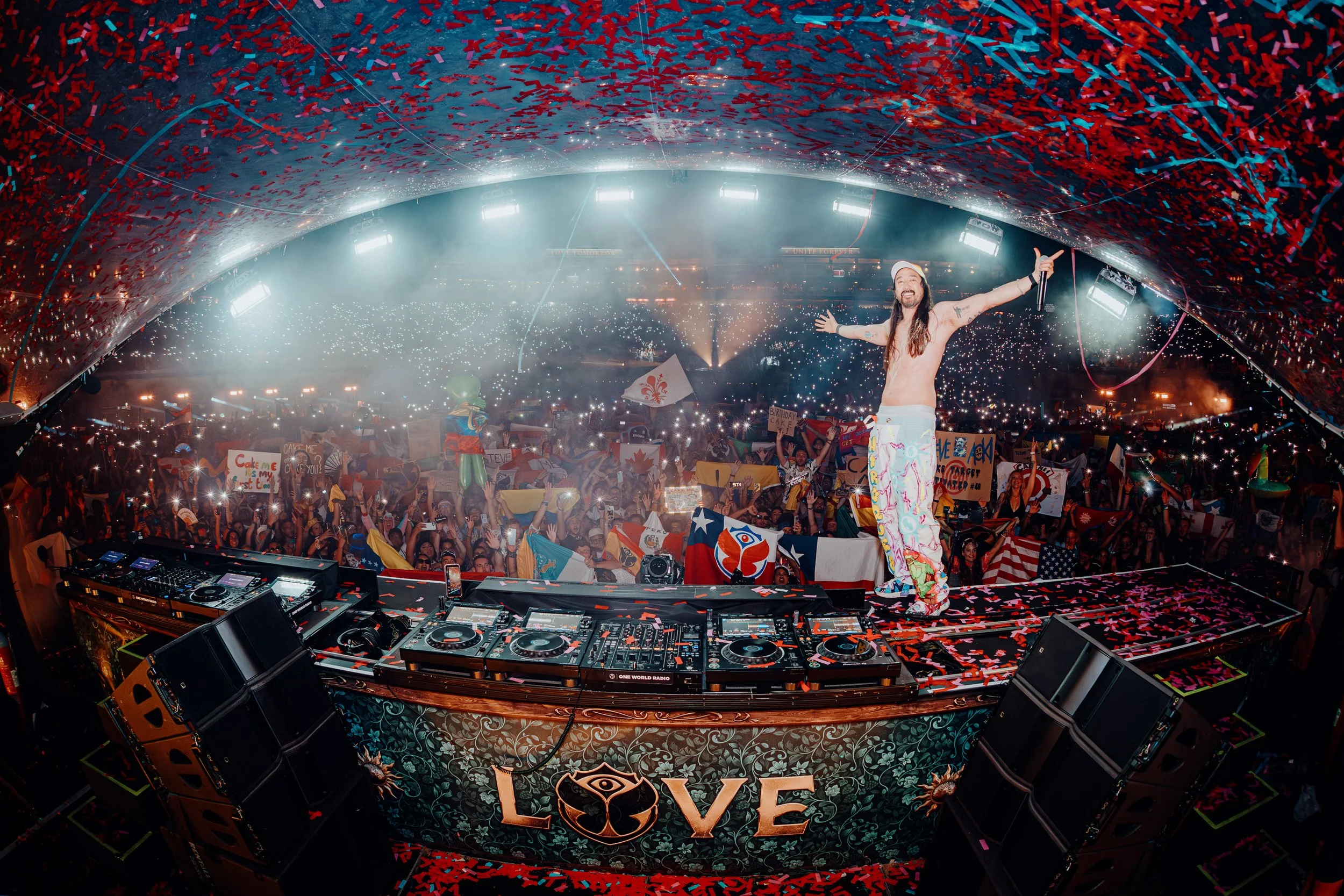 Steve Aoki Tomorrowland Group Photo standing on stage at a concert with arms raised, surrounded by a large crowd holding signs, flags, and lights, with confetti and bright lights overhead. Taken by James Bridle Music Photographer 2025